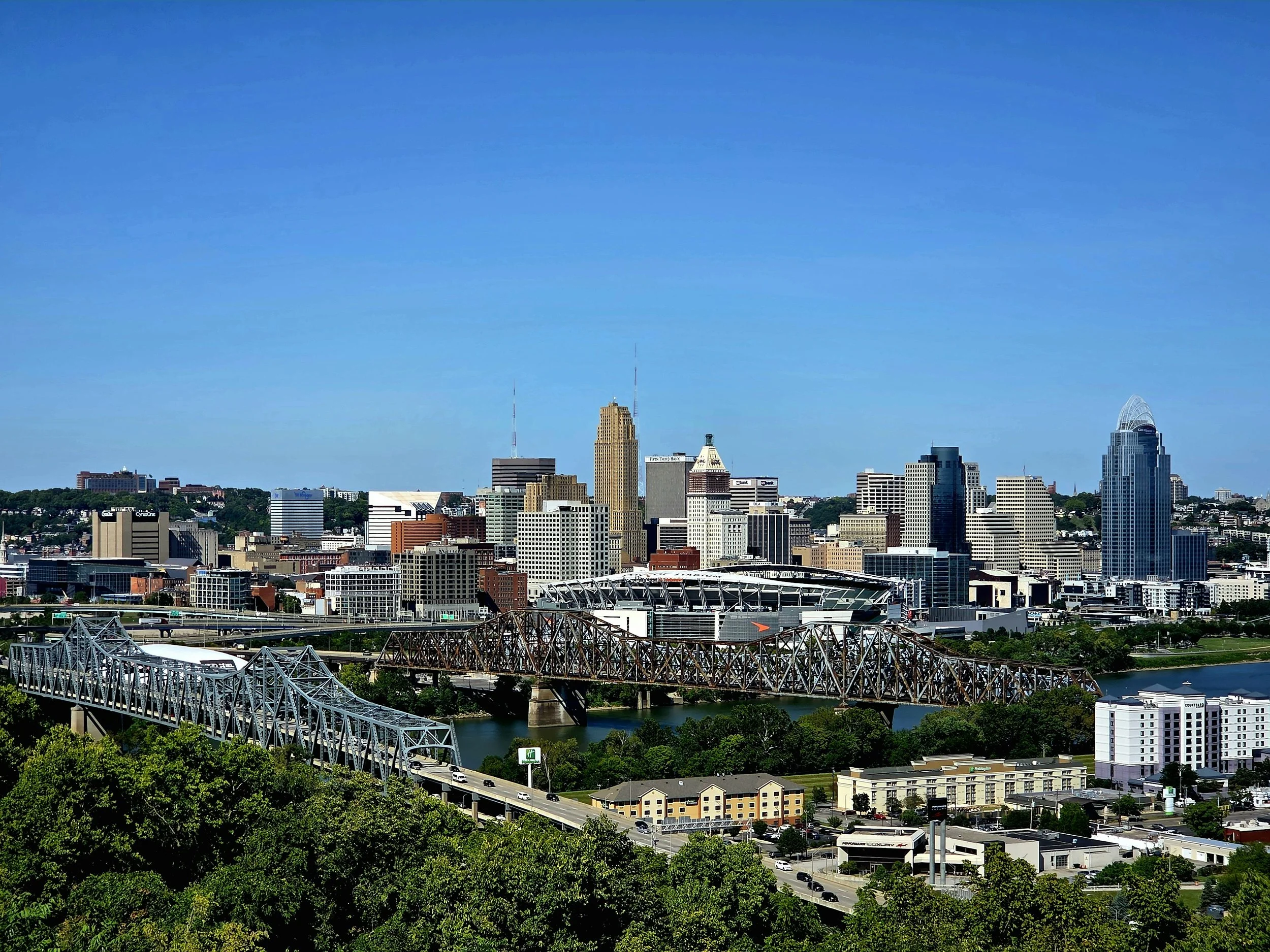 City skyline of downtown with several high-rise buildings, including one with a pointed top, and two radio towers, against a bright blue sky. A metal bridge crosses a river in the foreground.