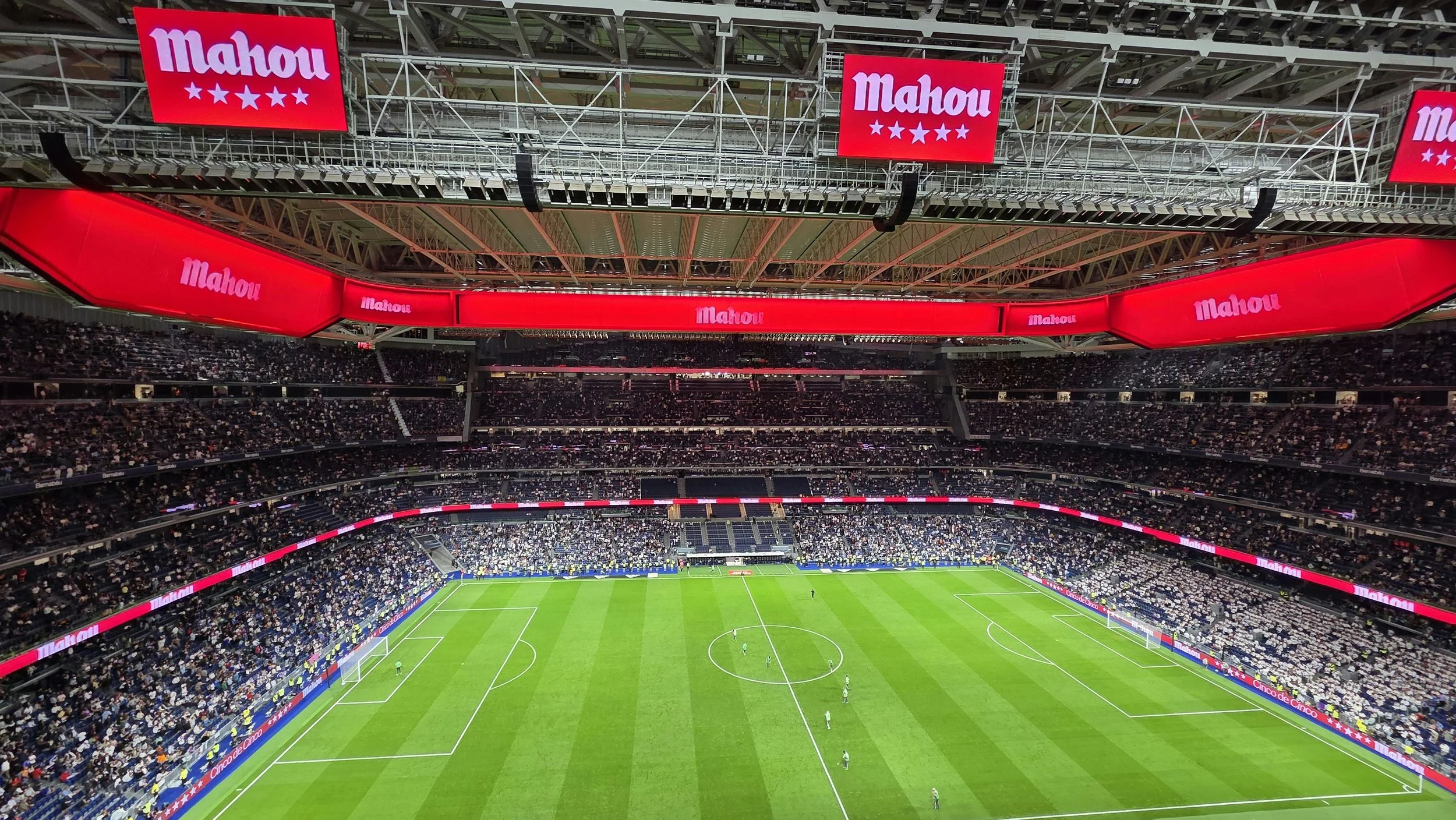 Interior view of a large soccer stadium filled with spectators, with digital advertising screens displaying the Mahou logo, and players visible on the field.