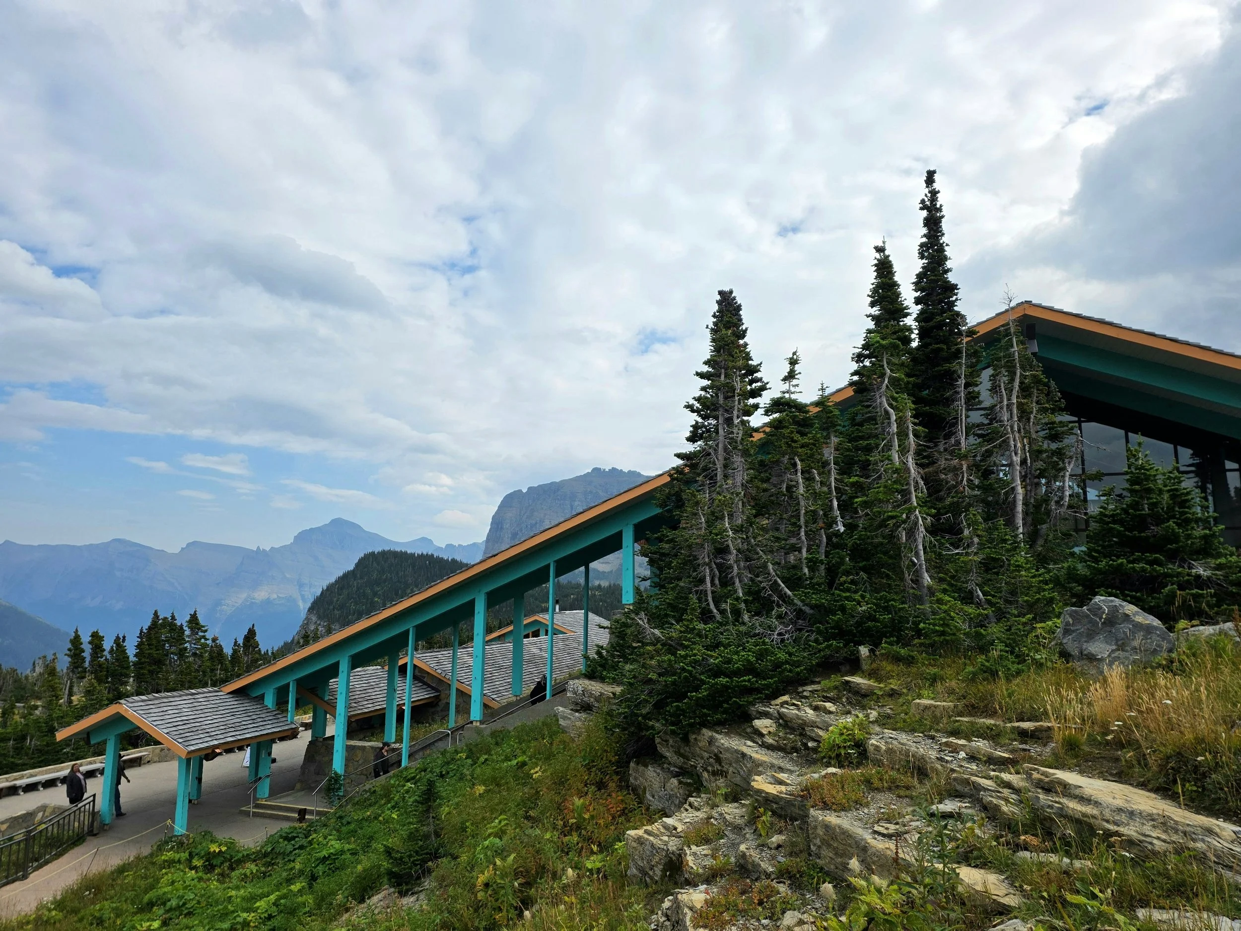 Mountain landscape with evergreen trees, a building with a colorful roof, and a cloudy sky.