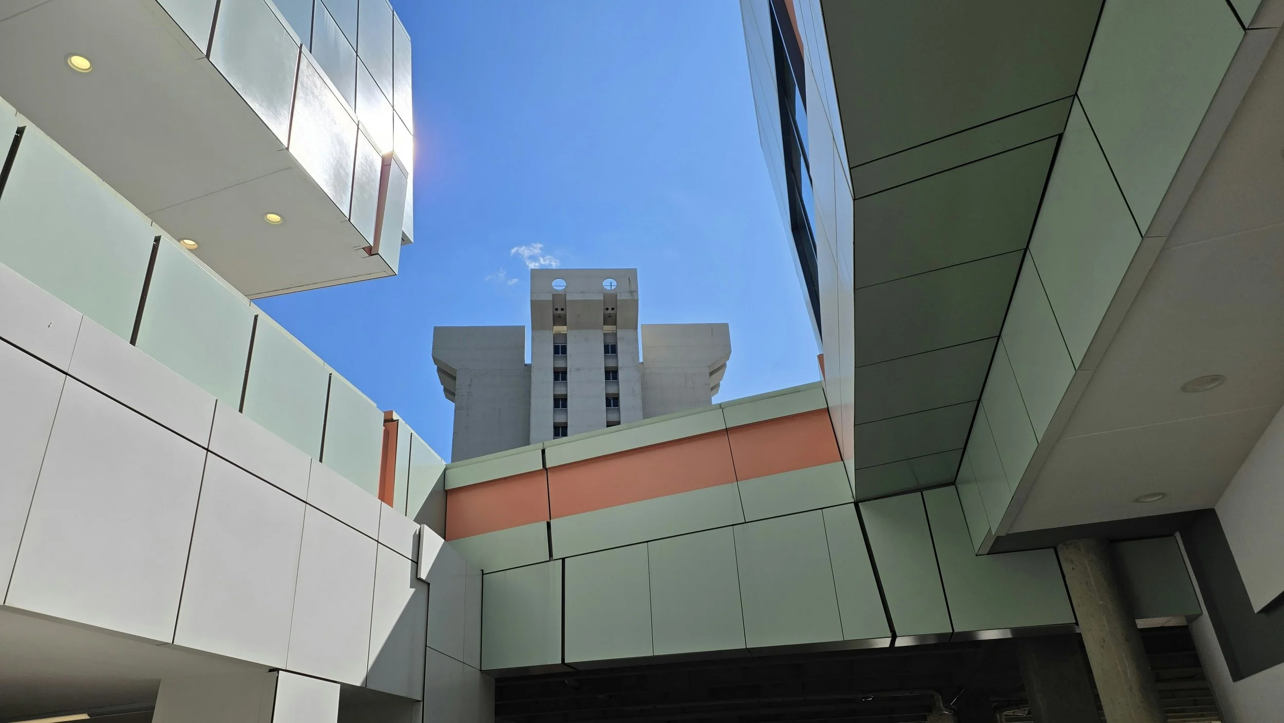 Looking up between two modern buildings with reflective glass windows and colorful panels, with a clear blue sky and a tall white building with unique architectural features visible in the background.