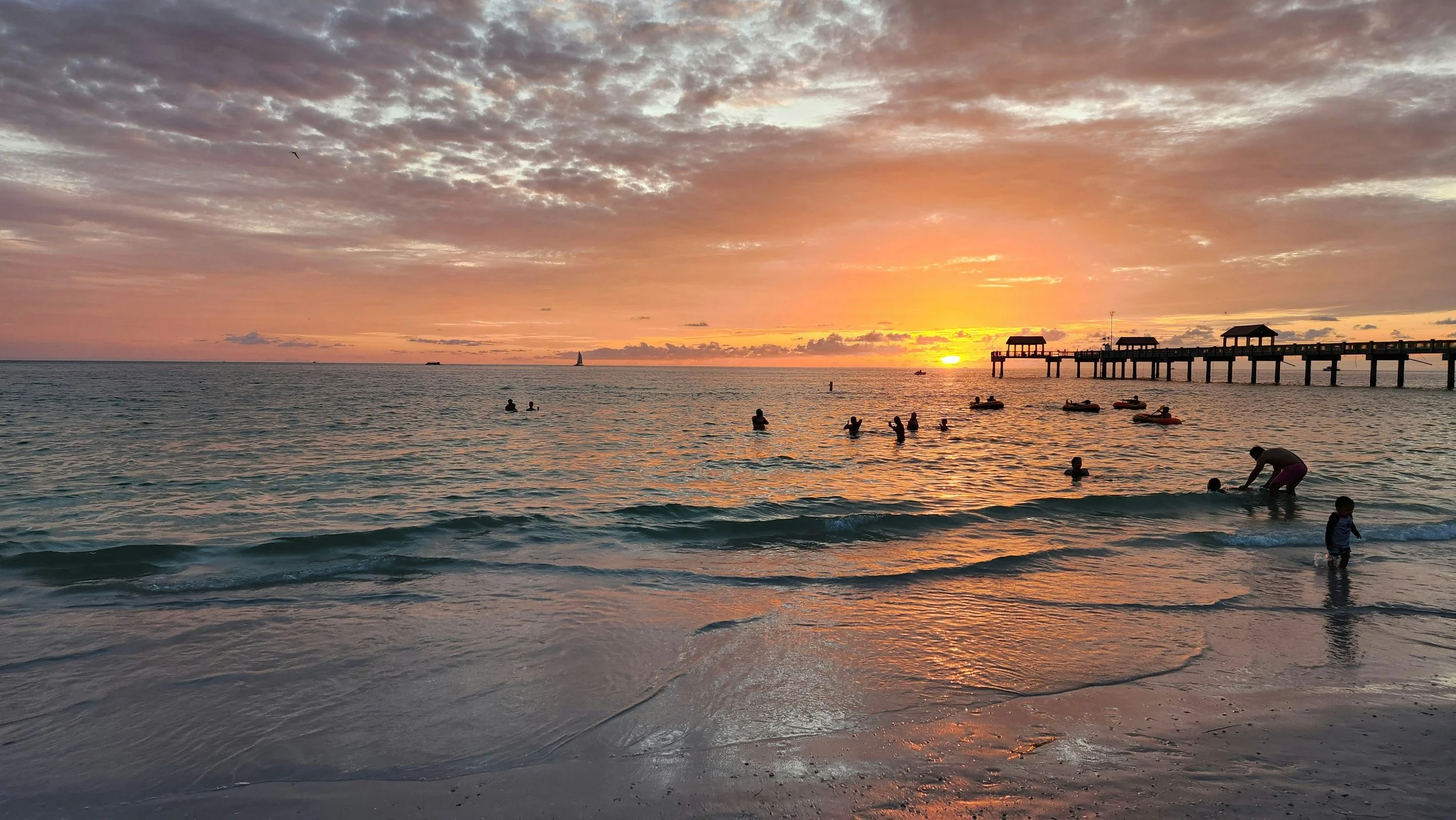 People swimming and playing in the ocean during a colorful sunset with a pier and sailboat in the background.
