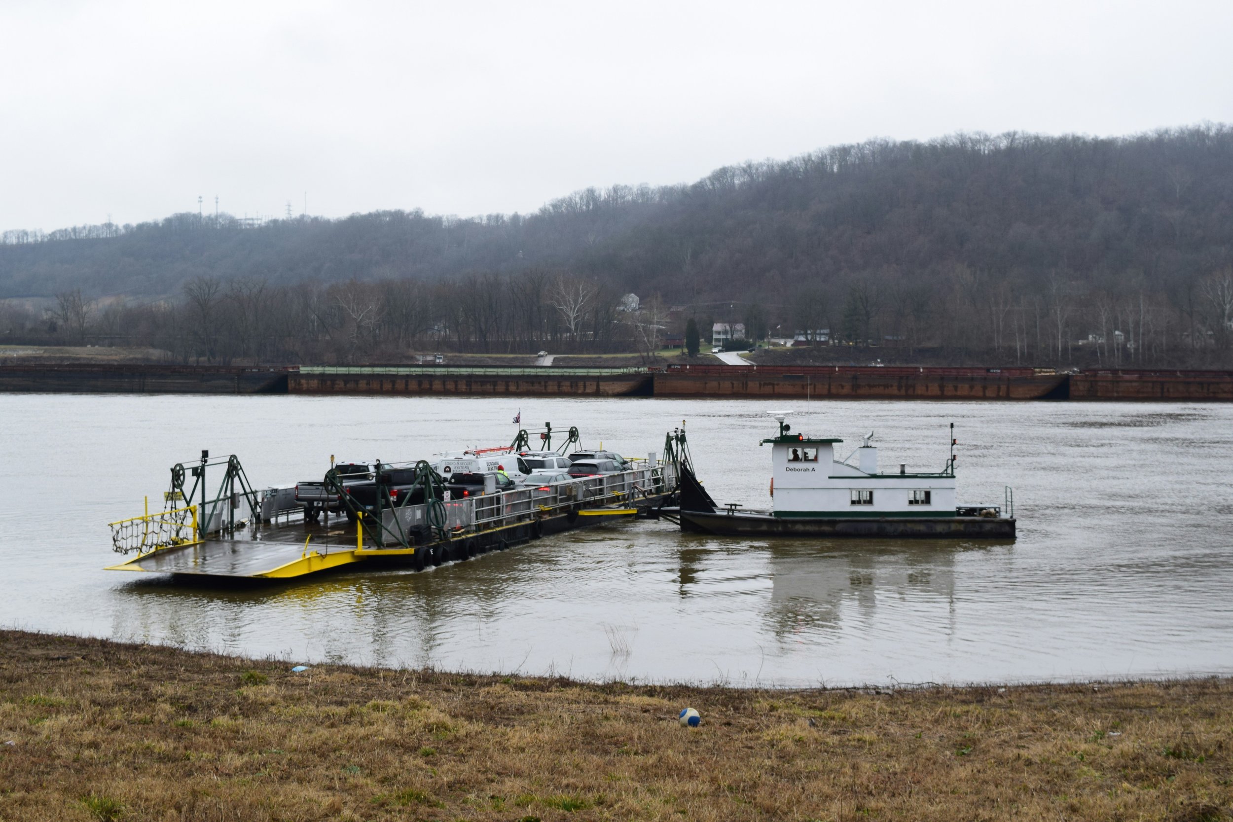 A ferry boat with cars on board sailing on a river, with a forested hill in the background and a cloudy sky.