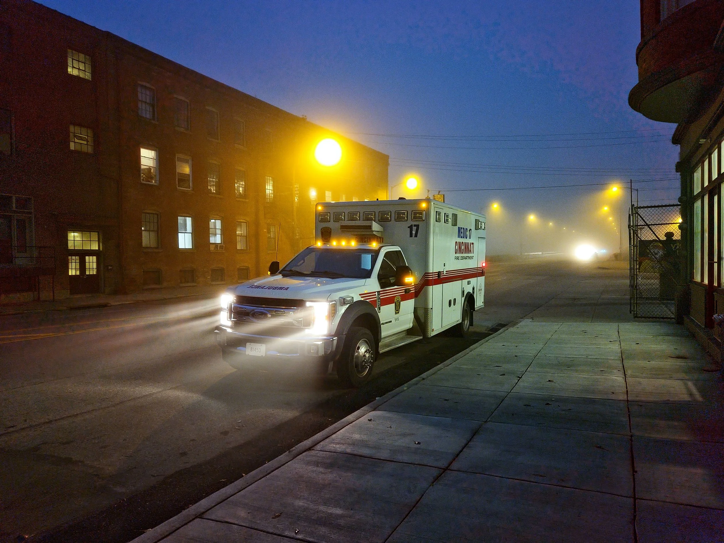Nighttime scene showing an emergency vehicle with flashing lights parked on a city street, with a building on the left and streetlights illuminating the foggy scene.