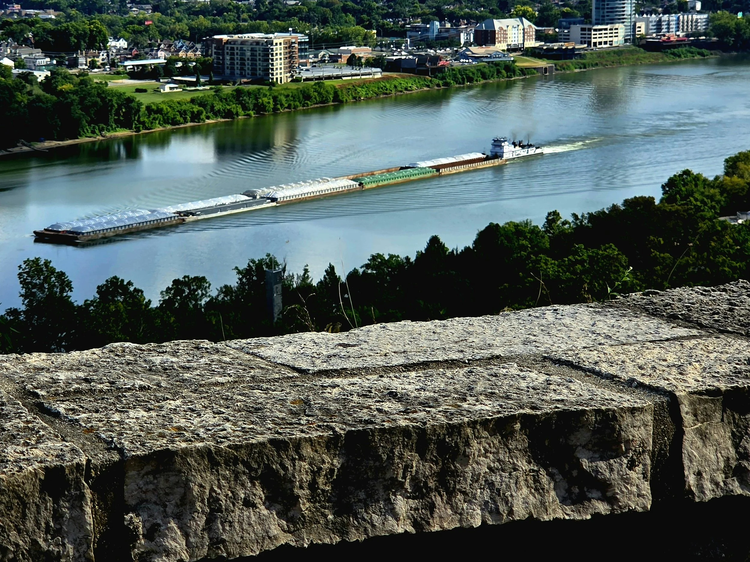 Aerial view of a river with a cargo boat moving through, with a cityscape of buildings and houses in the background and trees along the riverbanks.