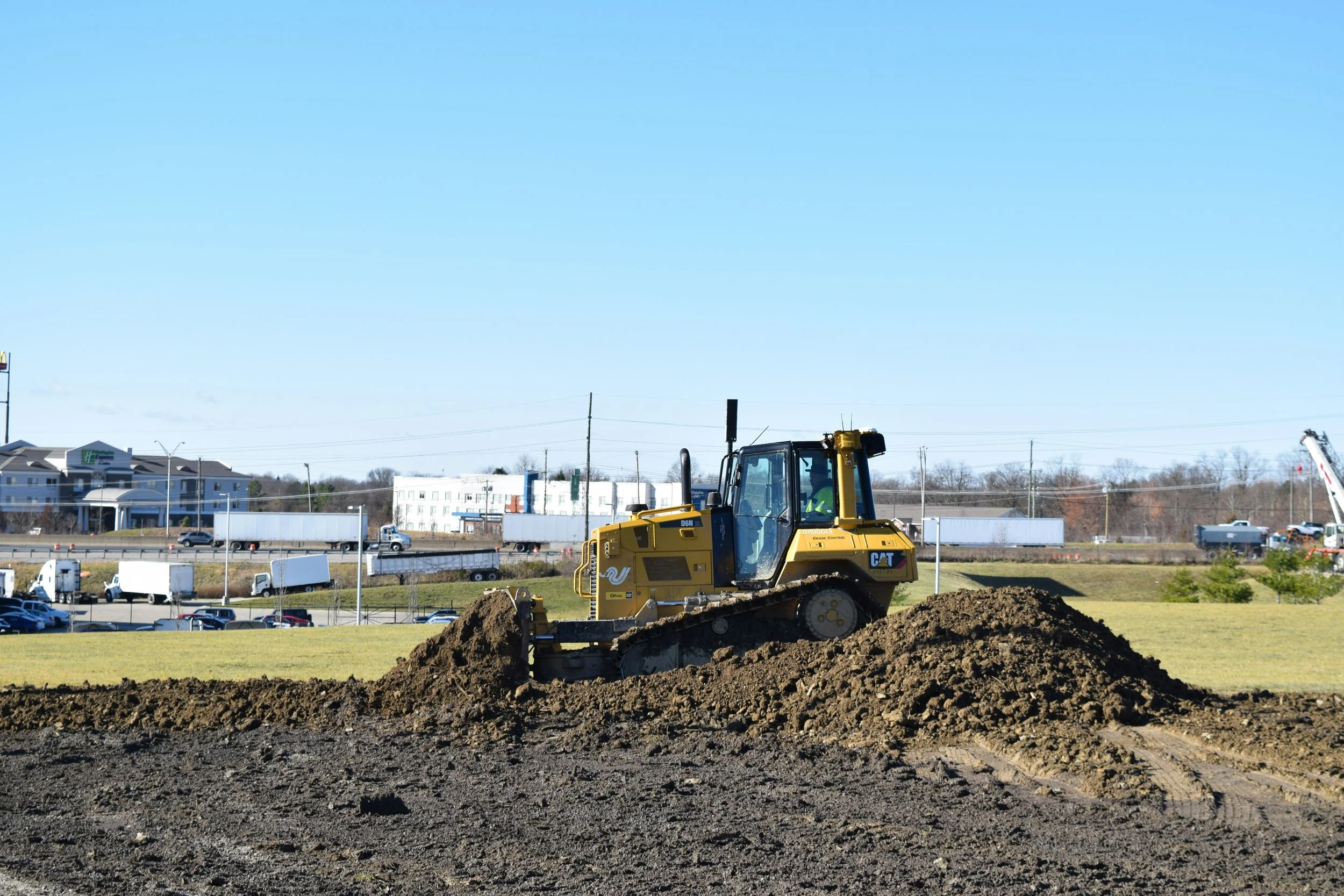 Construction site with a yellow bulldozer moving dirt, background shows buildings, cars, and power lines under a blue sky.