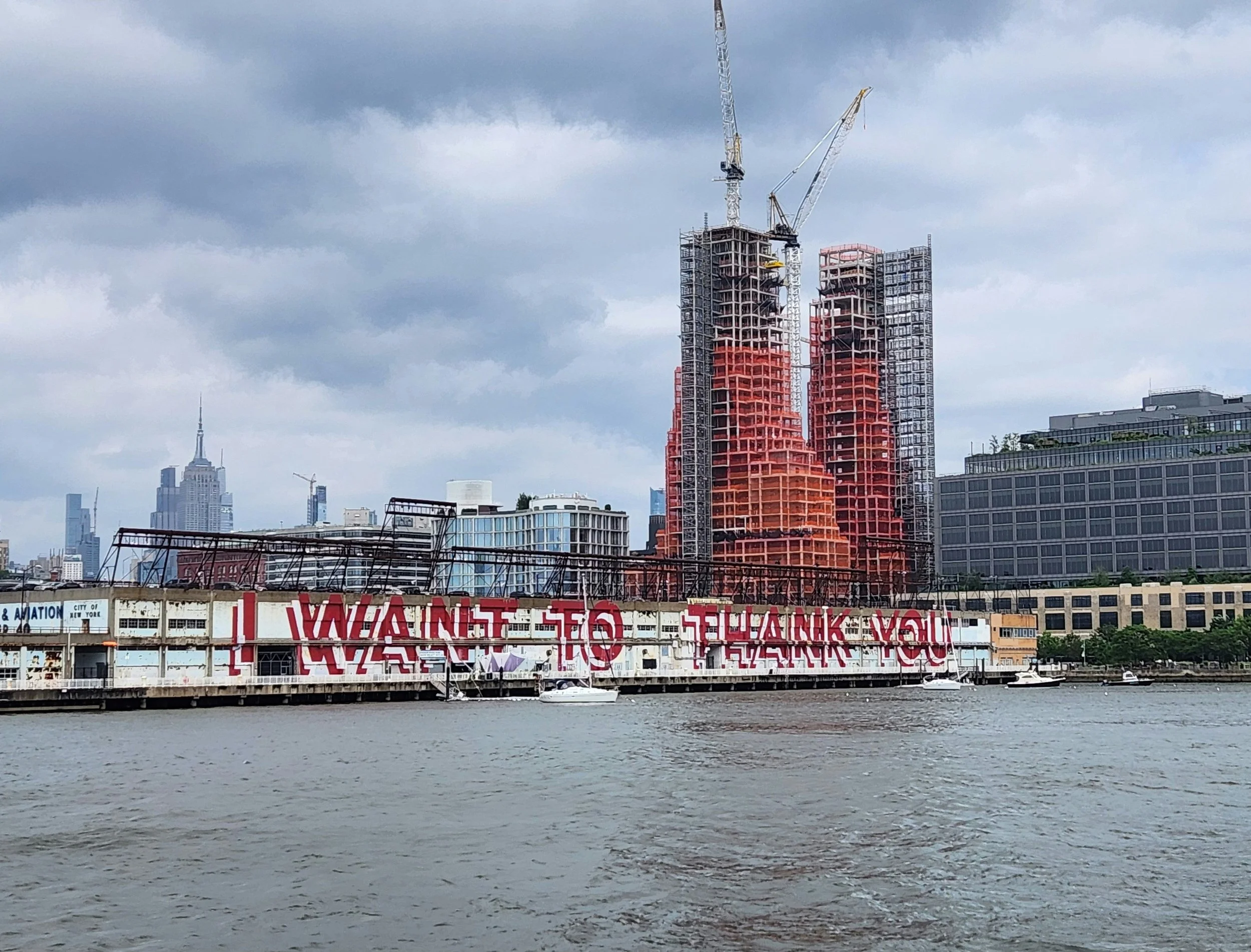 View of a city skyline from across a river with a boat in the foreground, and two high-rise buildings under construction with cranes. A large sign with partially visible red and white lettering is on a building along the riverbank.