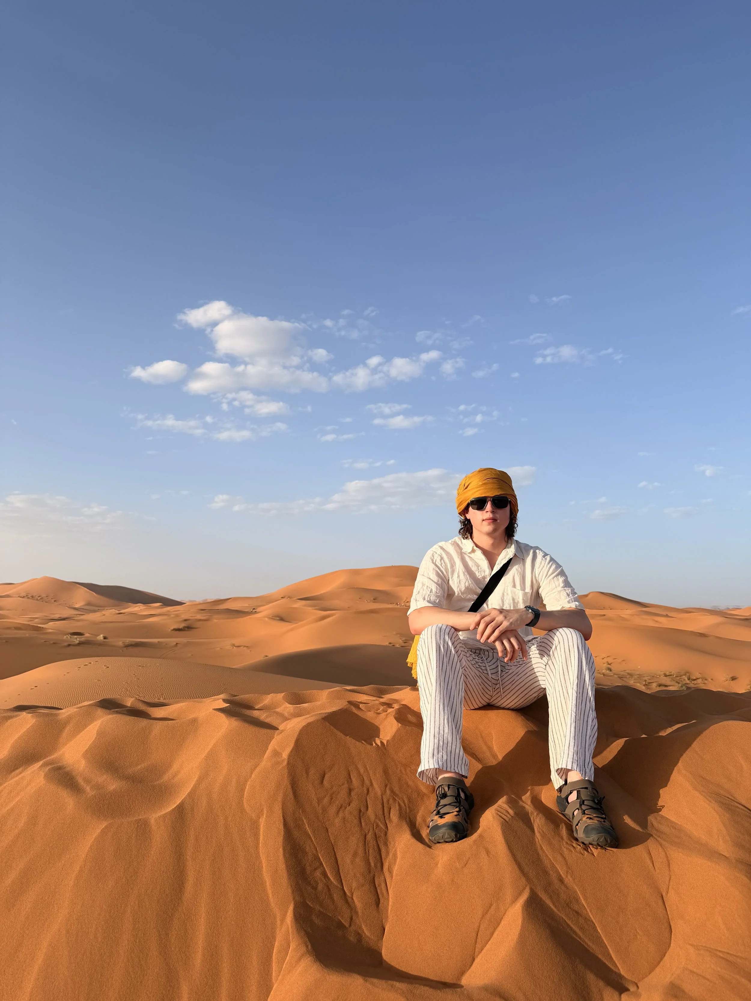 A person sitting on a sand dune in a desert landscape with a clear blue sky and a few scattered clouds, wearing sunglasses, a yellow headscarf, a white shirt, striped pants, and hiking sandals.