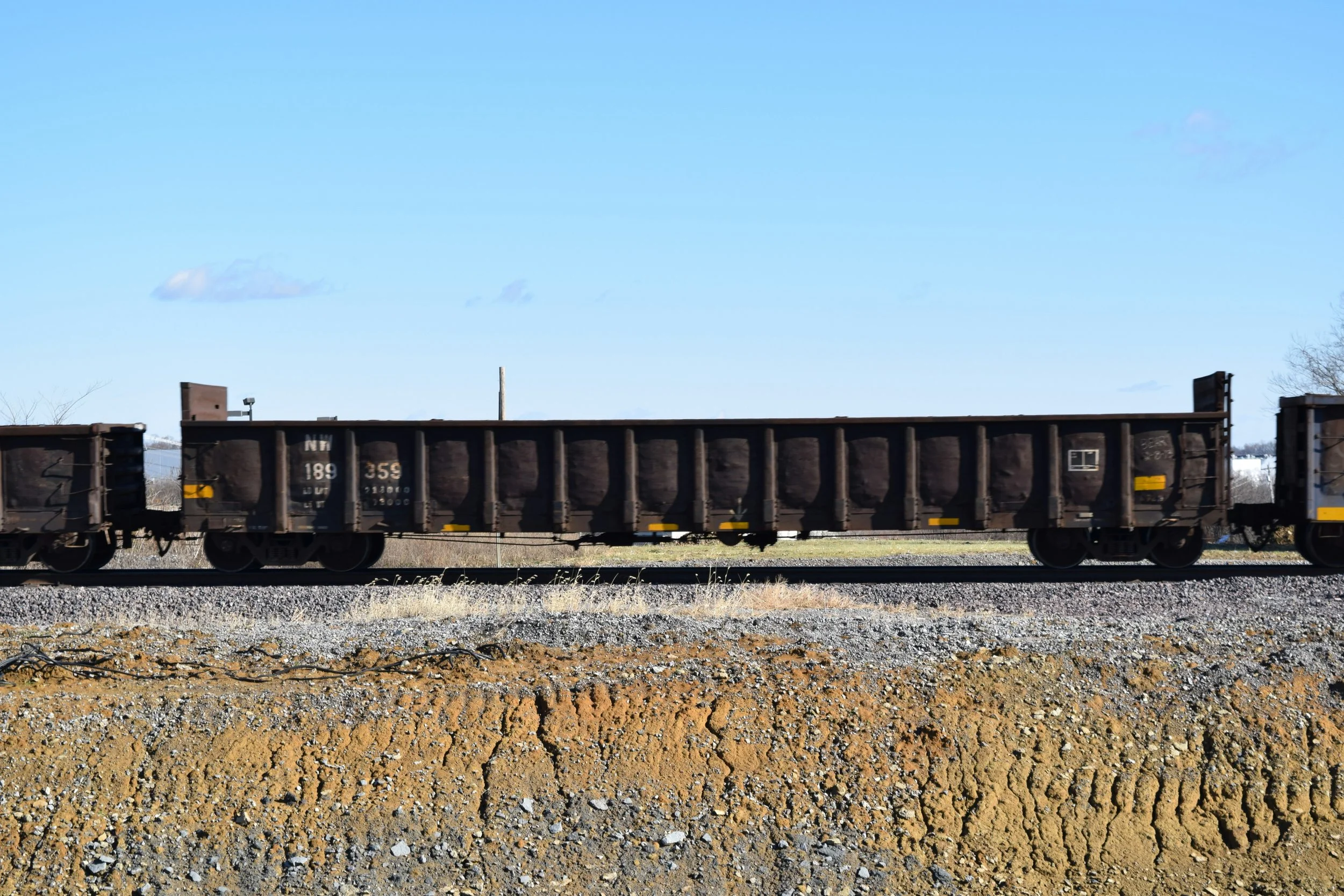 A photograph of a freight train with black cargo cars on train tracks, with a gravel and dirt ground in the foreground and a clear blue sky with a few scattered clouds in the background.