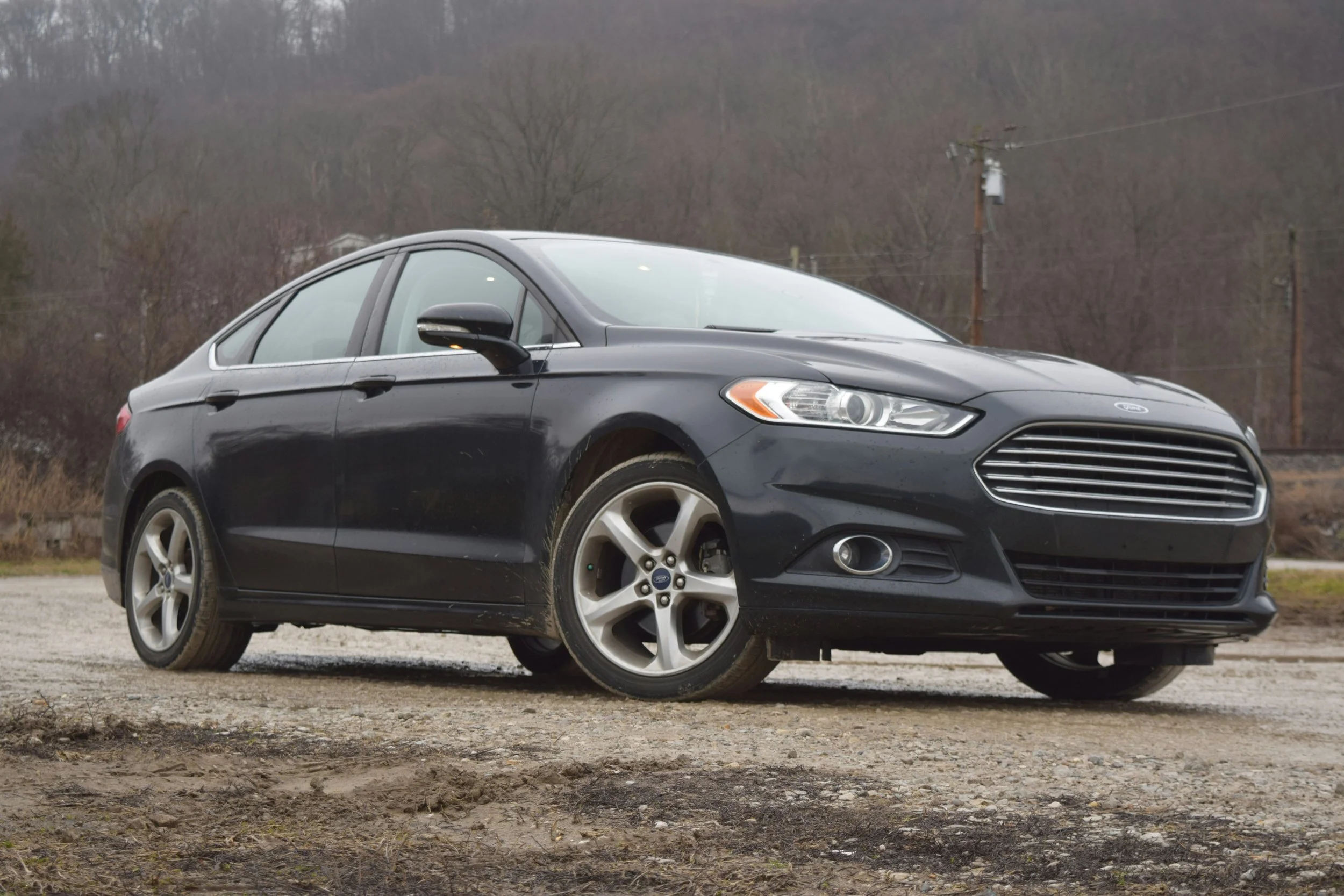Black sedan parked outdoors on a gravel surface with a background of leafless trees and utility poles under a cloudy sky.