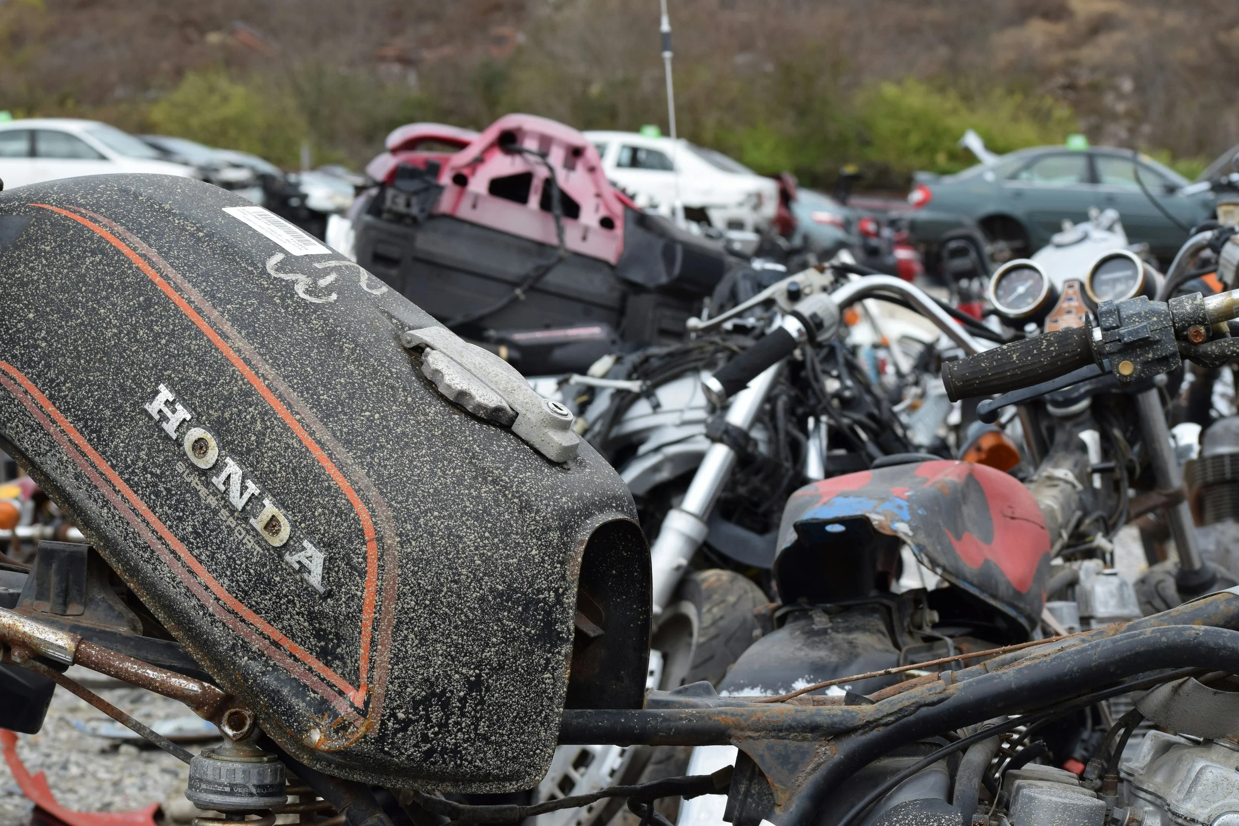 A close-up of a dusty, damaged motorcycle with a Honda logo, with a heap of wrecked cars and vehicles in the background.