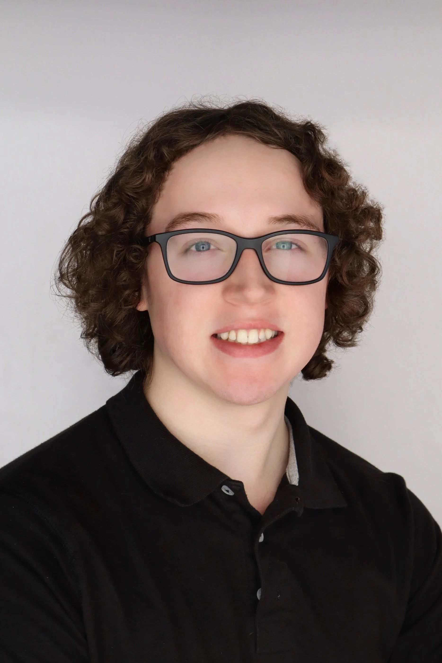 A young person with curly hair and glasses smiling, wearing a black polo shirt, against a plain light background.