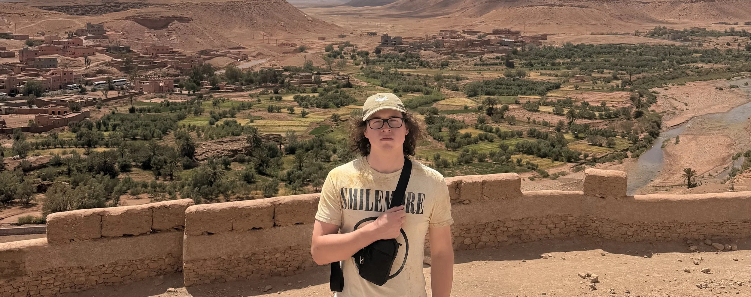 Young man standing on a stone wall, wearing glasses, a beige cap, and a light-colored t-shirt with the word 'SMILEMORE' printed on it. The background features an arid landscape with a river, green fields, trees, and a small town with buildings, set in a mountainous desert region.