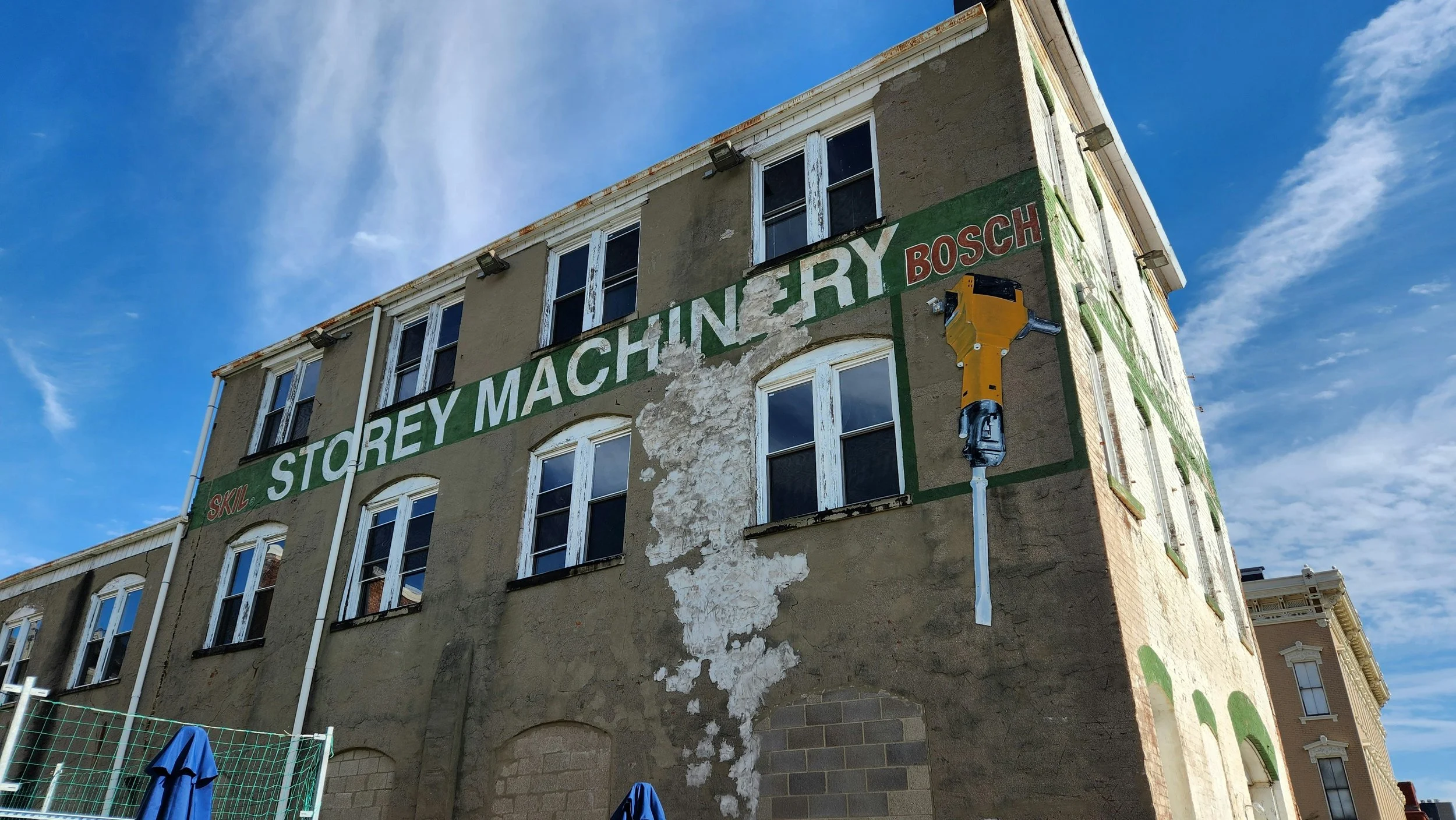 Old building with a sign that reads 'Storey Machinery Bosch' with some peeling paint and a large yellow power tool attached to the wall, under a blue sky with some clouds.