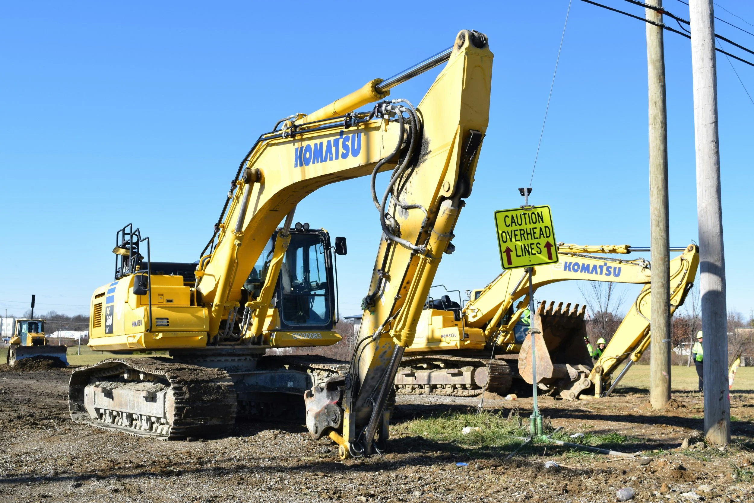 Yellow Komatsu excavator at a construction site with a caution sign for overhead lines and workers in safety gear in the background.