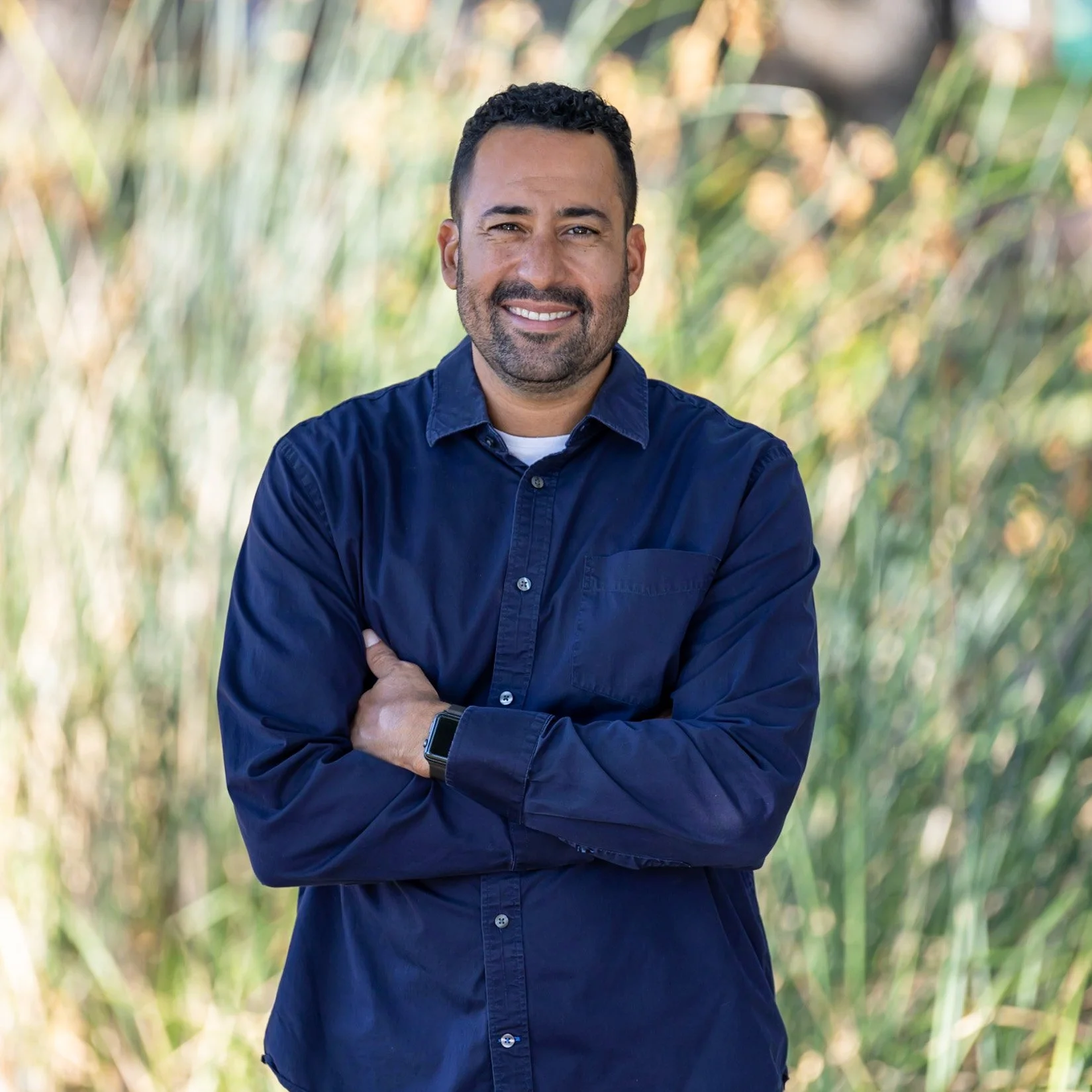 A man with short curly dark hair and a beard, smiling, wearing a navy blue button-up shirt and a smartwatch, standing outdoors with blurred greenery in the background.
