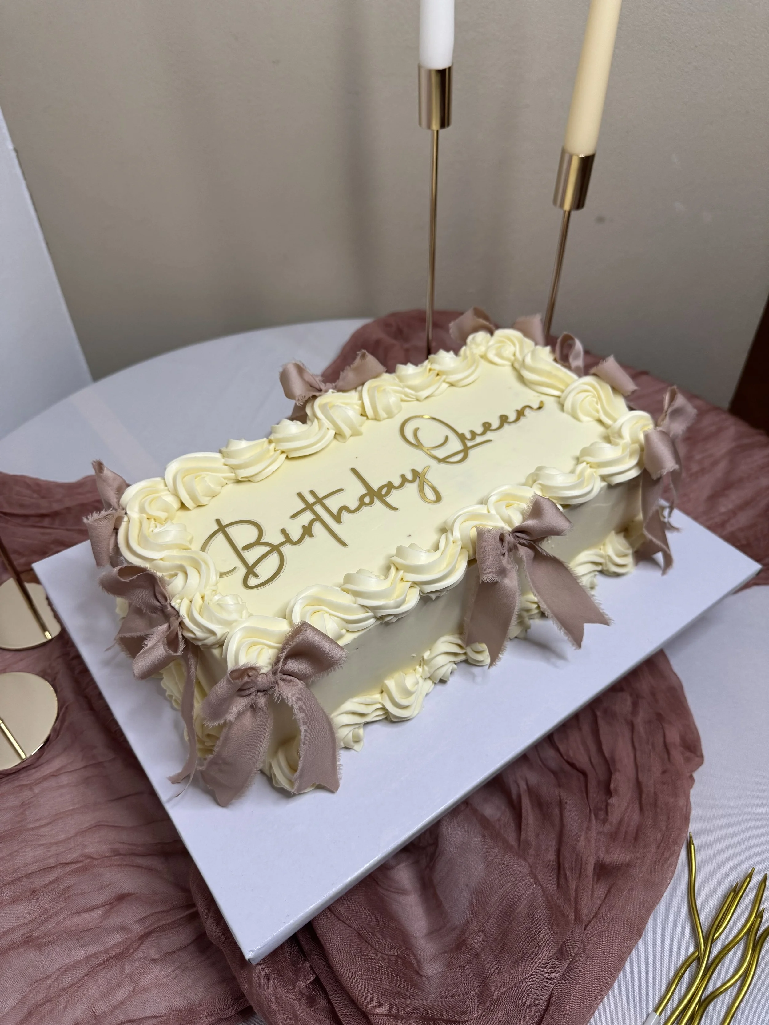 Gâteau d'anniversaire en forme d'Italian Cake avec décoration en crème et rubans en satin, inscription #Happy Queen, sur une table avec des décorations roses et or. 