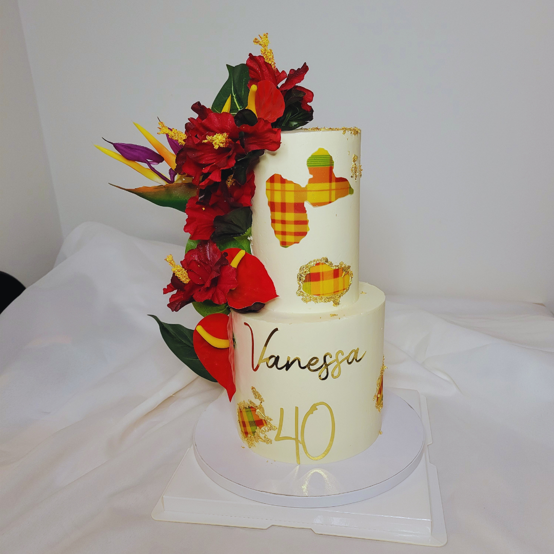 Gâteau blanc à deux niveaux décoré avec des fleurs rouges, vertes, jaunes et violettes, avec des feuilles vertes, et des décorations en papier avec un motif à carreaux en jaune, rouge et orange. Le gâteau a le nom 'Vanessa' écrit en or et le chiffre 