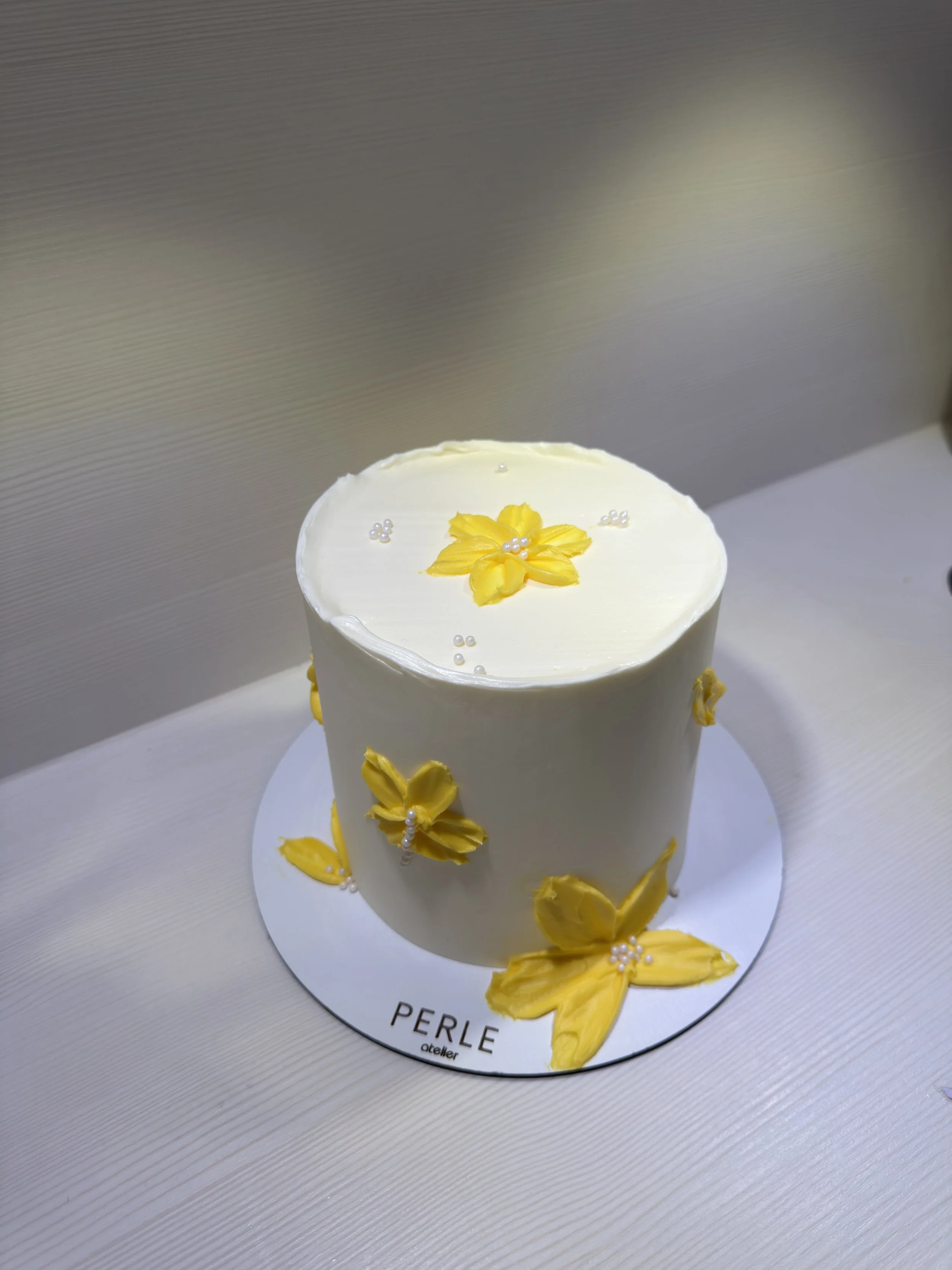 Gâteau blanc décoré avec des fleurs jaunes en crème au beurre et de petites perles. Le gâteau est placé sur un support blanc avec l'inscription 'PERLE atelier'