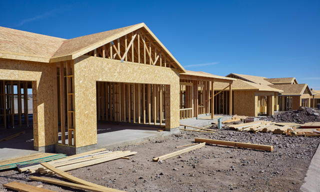 New residential houses under construction with wooden frames and unfinished exteriors, construction materials and tools on the ground, under a clear blue sky.
