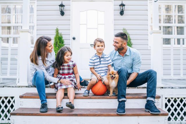 A family of four and a dog sitting on front porch steps in front of a white house, smiling and enjoying each other's company.