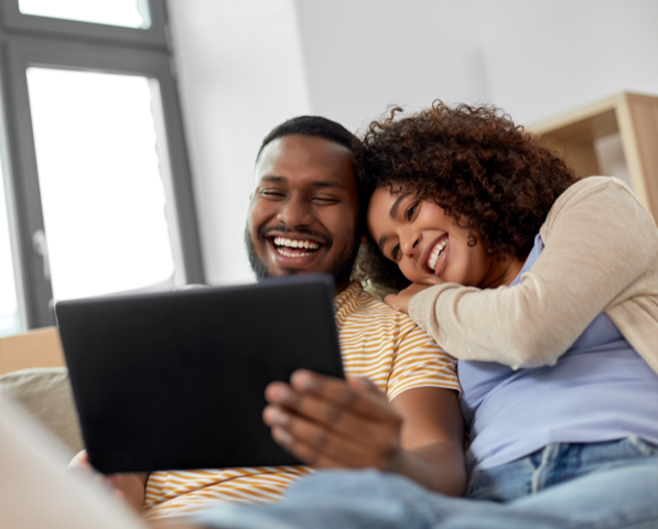 A happy couple sitting close together on a couch, looking at a tablet and smiling.