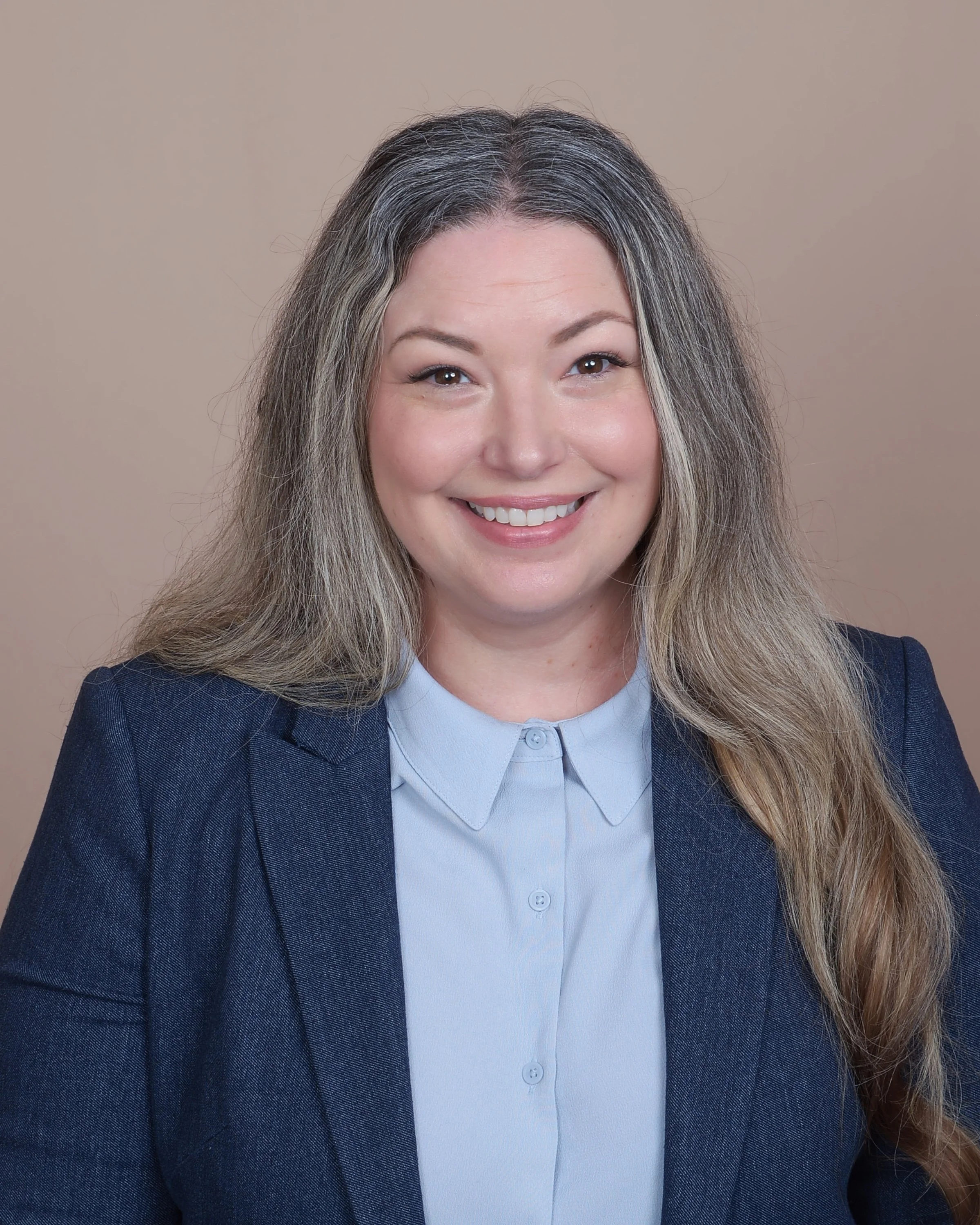 Smiling woman with long, wavy gray and blonde hair wearing a navy blazer and light blue shirt, against a beige background.