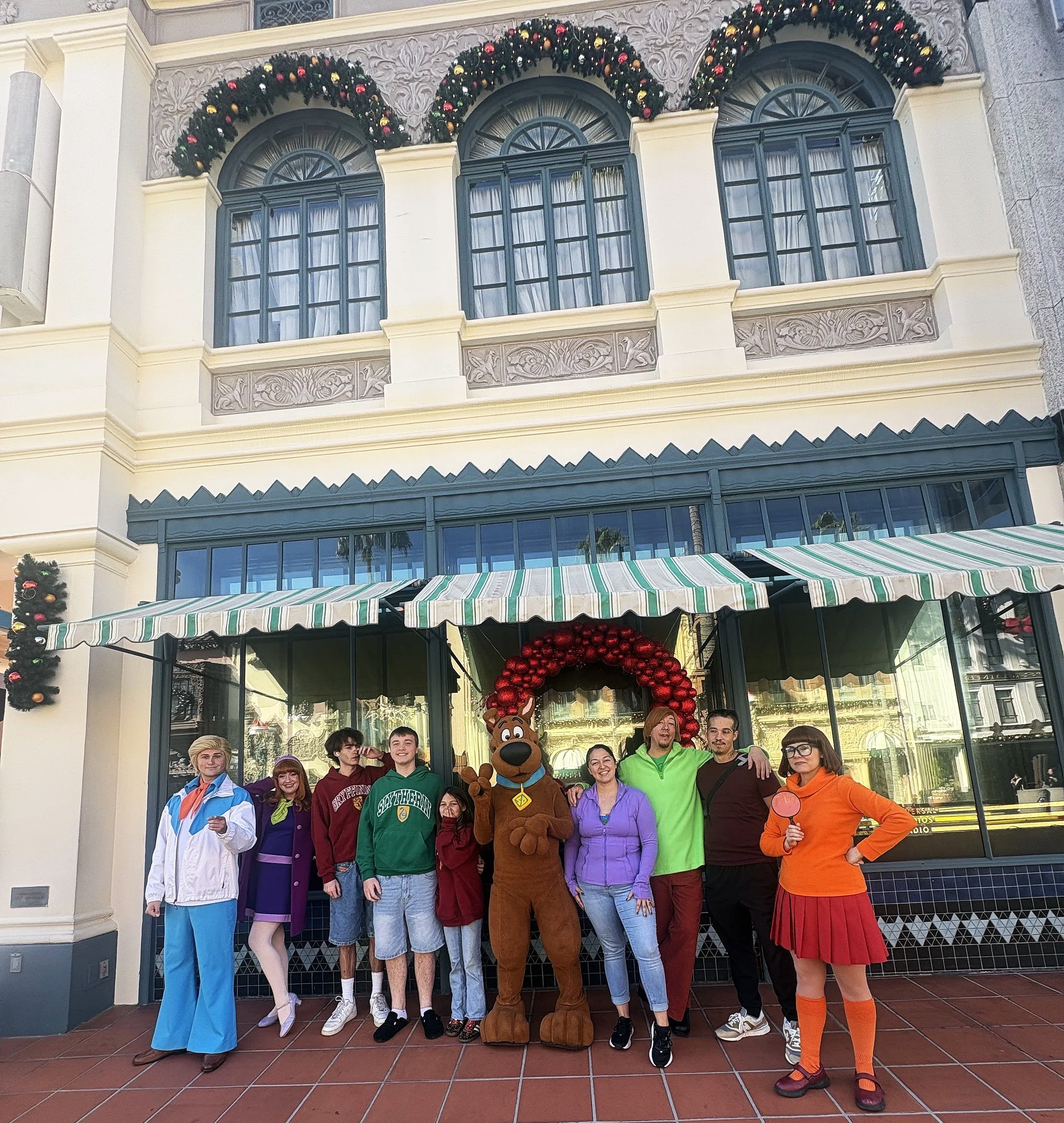 Group of people in colorful costumes and casual clothing posing in front of a building decorated for Christmas, including a person in a Scooby-Doo costume and a woman in a red skirt and orange top, with Christmas wreaths and decorations visible.
