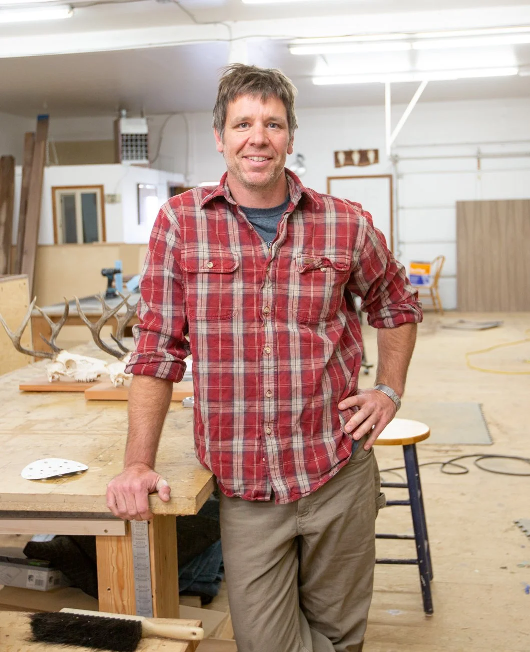 A man in a red plaid shirt leaning on a woodworking workbench in a workshop with tools and wood pieces in the background.