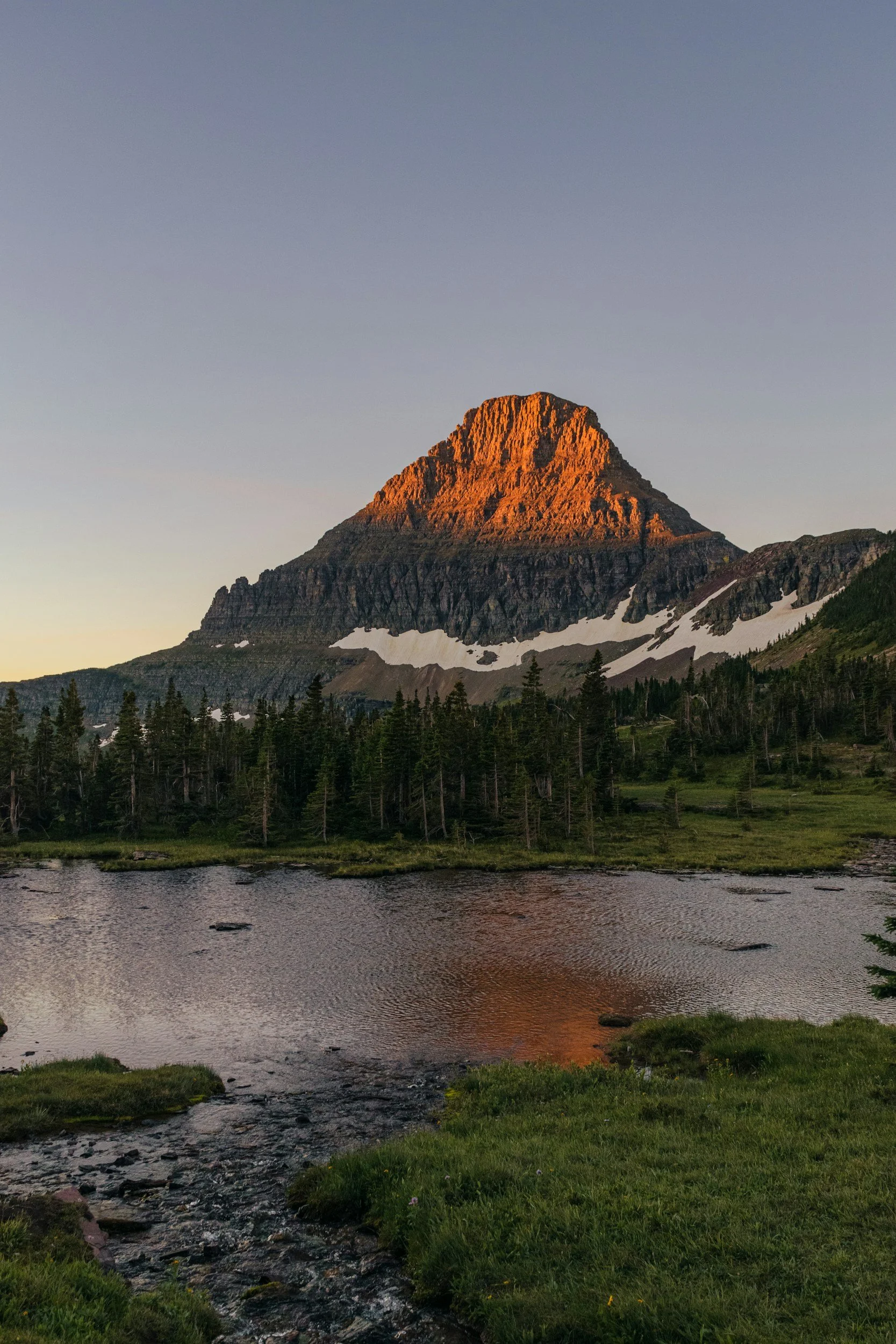 A mountain at sunset with a partly snow-covered lower slope, reflected in a calm river in the foreground, and a green grassy area with small yellow flowers along the river bank.