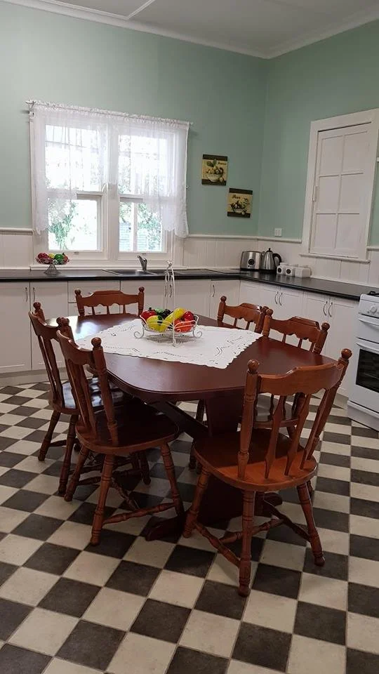A kitchen with a round wooden dining table surrounded by six matching wooden chairs on a black and white checkered floor. The table has a white lace tablecloth and a bowl of colorful plastic eggs. The background features white cabinets, a window with white lace curtains, and wall decorations.