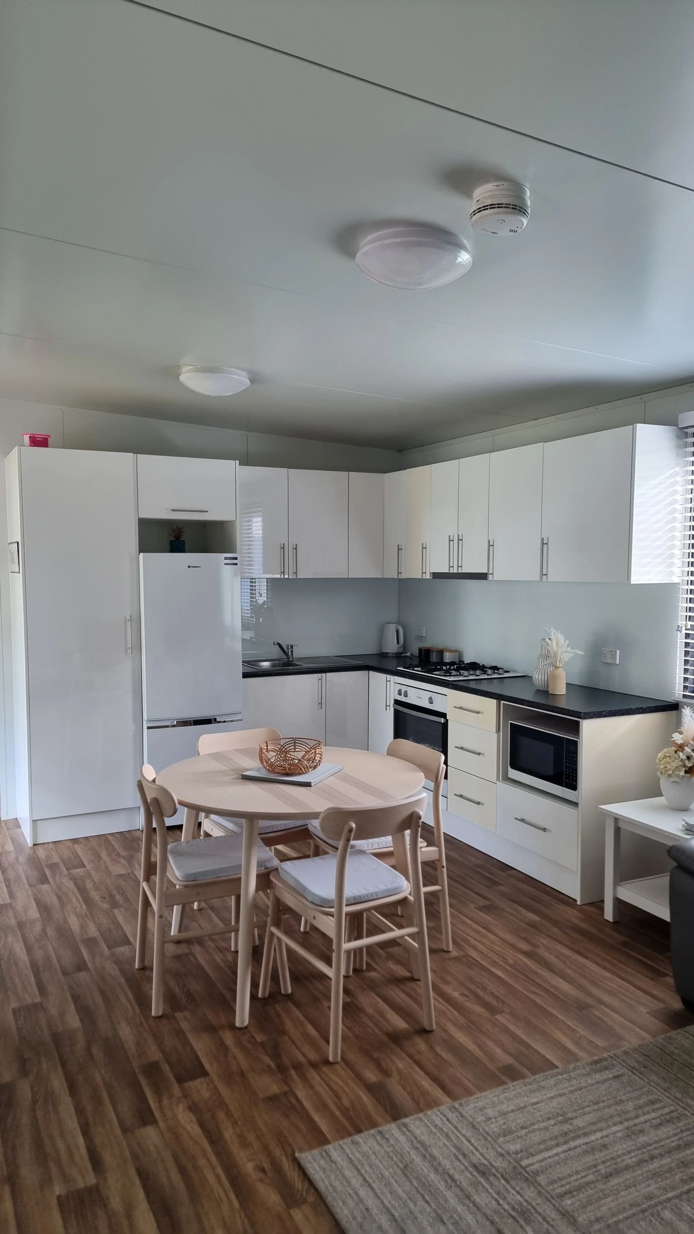 A modern kitchen with white cabinets, a white refrigerator, and a small dining table with four chairs. There are kitchen appliances on the countertop, a small decorative plant on the table, and a window with blinds on the right side. The room has woo
