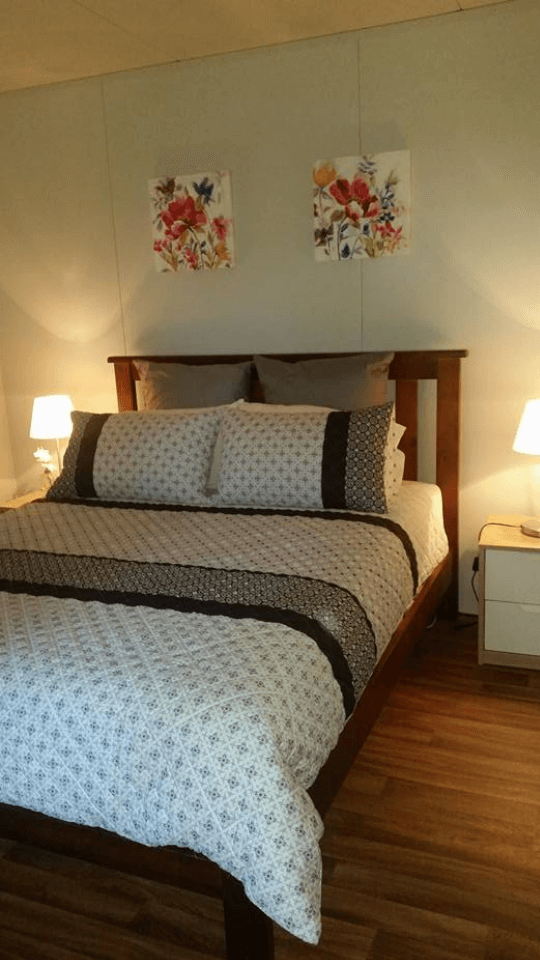 A bedroom with a wooden bed frame, two pillows with patterned pillowcases, two grey pillows, two nightstands with white lamps, and two floral paintings on the wall.