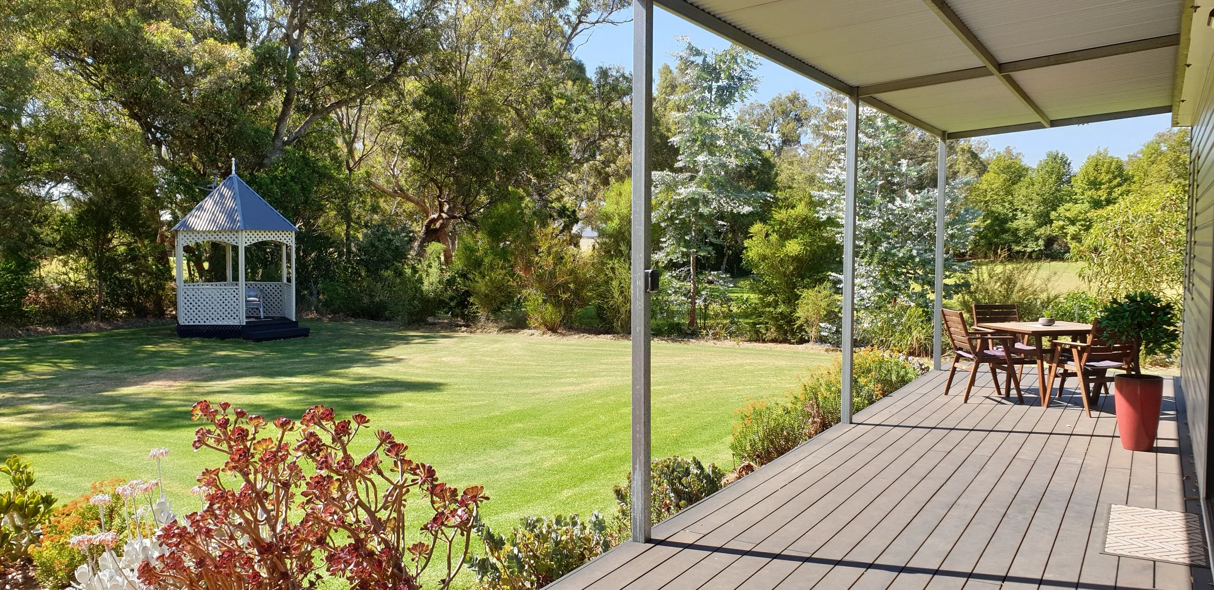 Backyard view from a deck with wooden chairs and a table, lush green lawn with trees, a white gazebo, and garden plants in the background, under a clear blue sky.