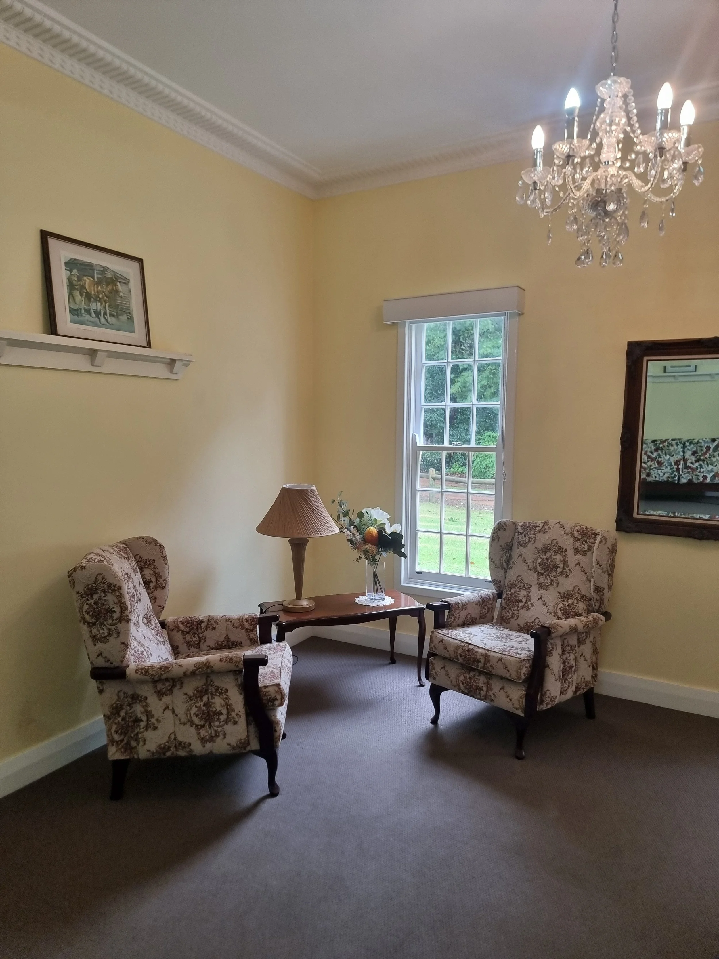 Living room with two upholstered armchairs with floral pattern, a wooden side table with a lamp and vase of flowers, a window with greenery outside, a framed mirror on the wall, and a chandelier hanging from the ceiling.