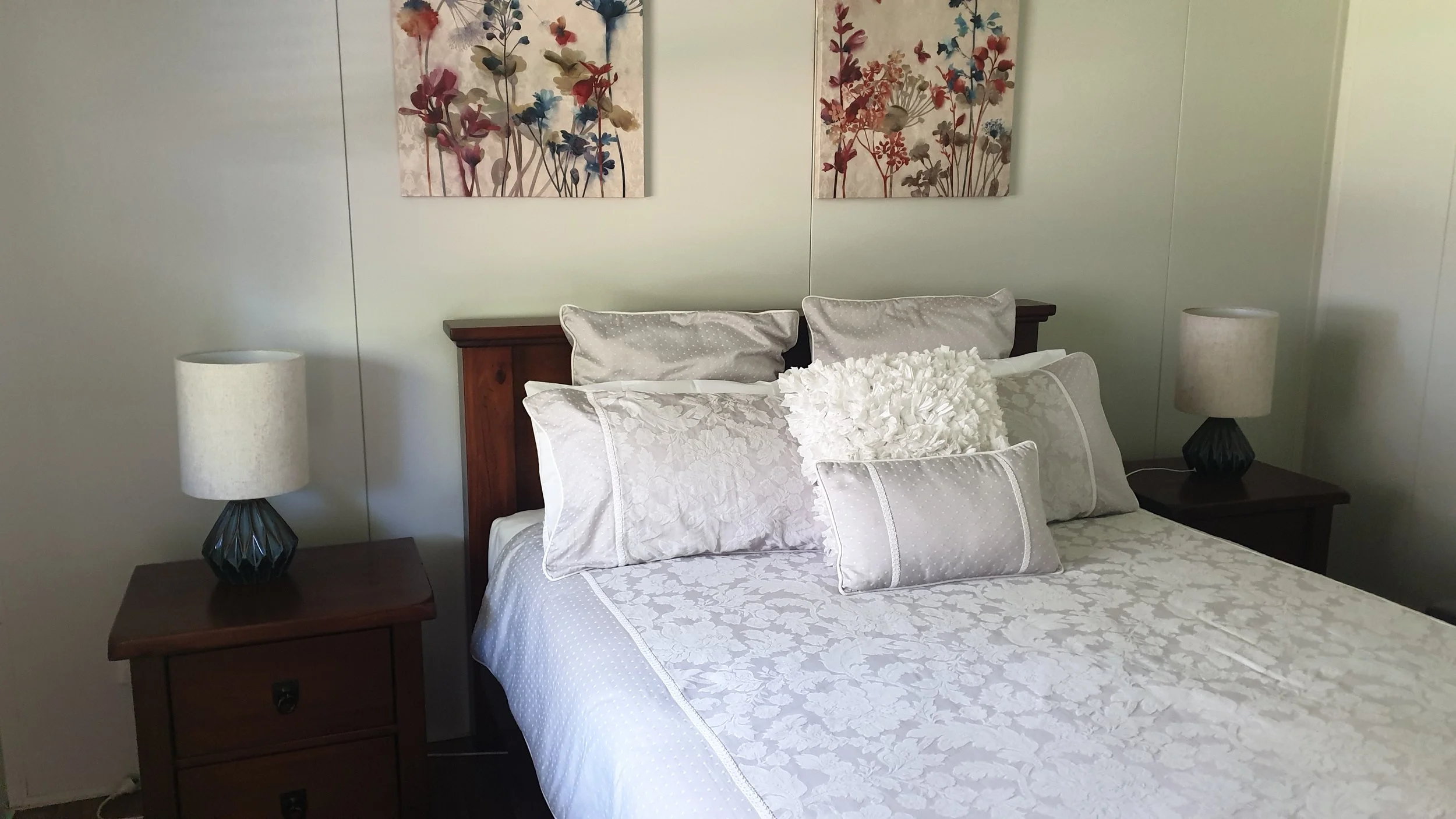 A bedroom with a bed, pillows, and floral wall art, flanked by two nightstands with lamps.