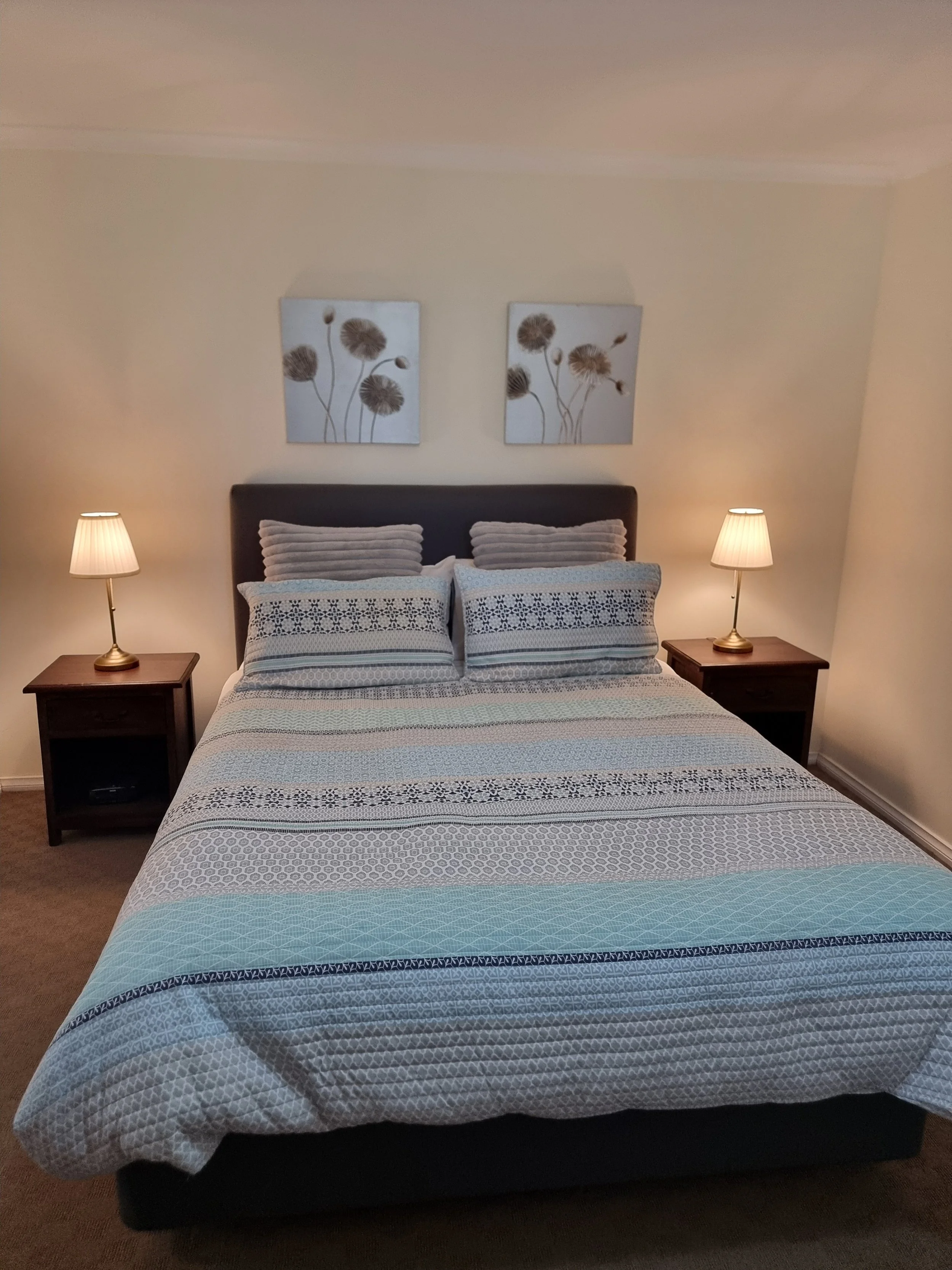 A neatly made bed with a dark upholstered headboard, patterned pillows, and a quilted bedspread in shades of blue, gray, and white. There are two matching nightstands with lamps on each side of the bed, three framed floral art pieces hang above the h