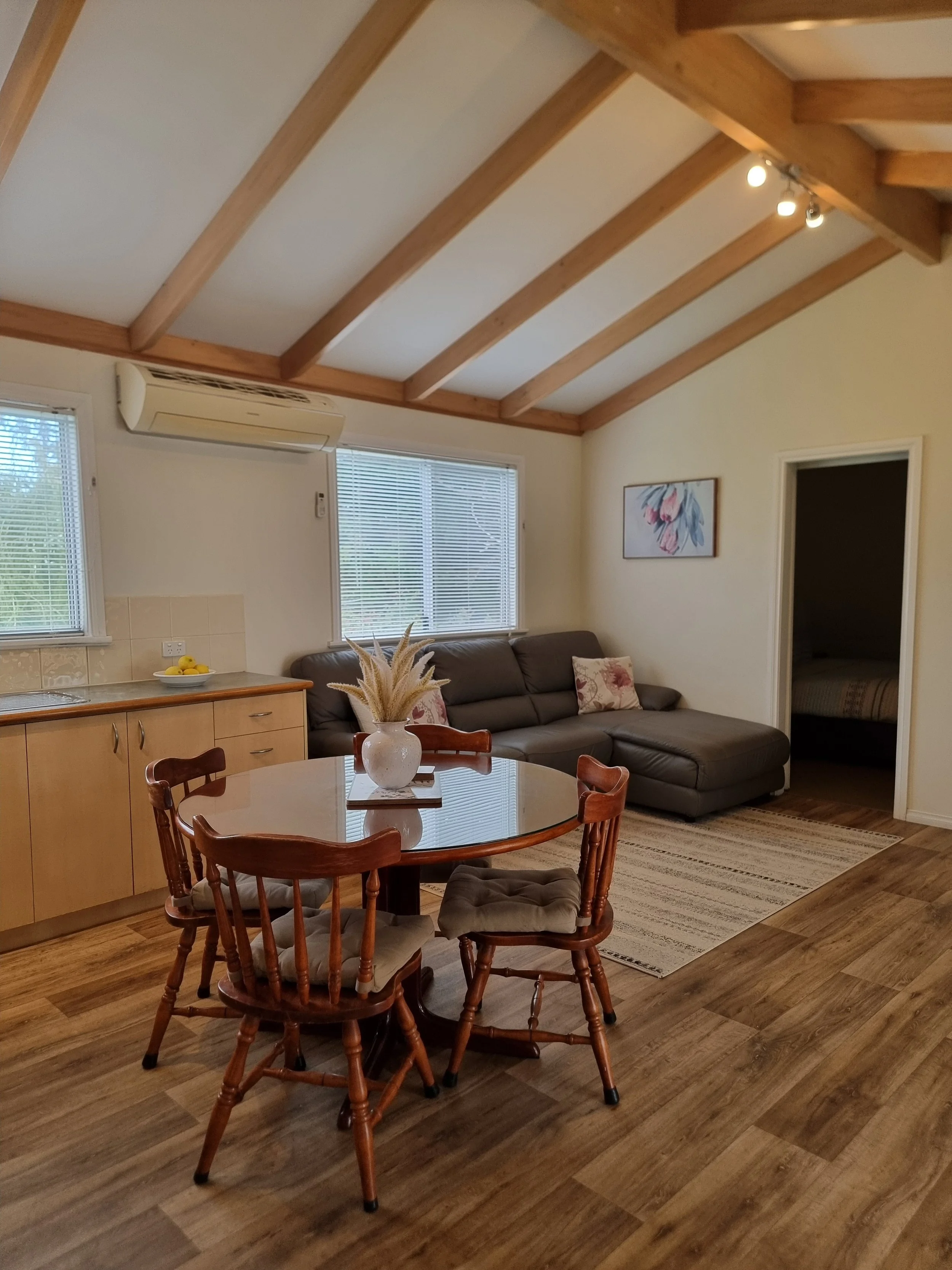 Living room with wood flooring, a round dining table with four wooden chairs, a beige sectional couch with floral pillows, a wall art painting, windows with blinds, an air conditioning unit, and a door leading to a bedroom.