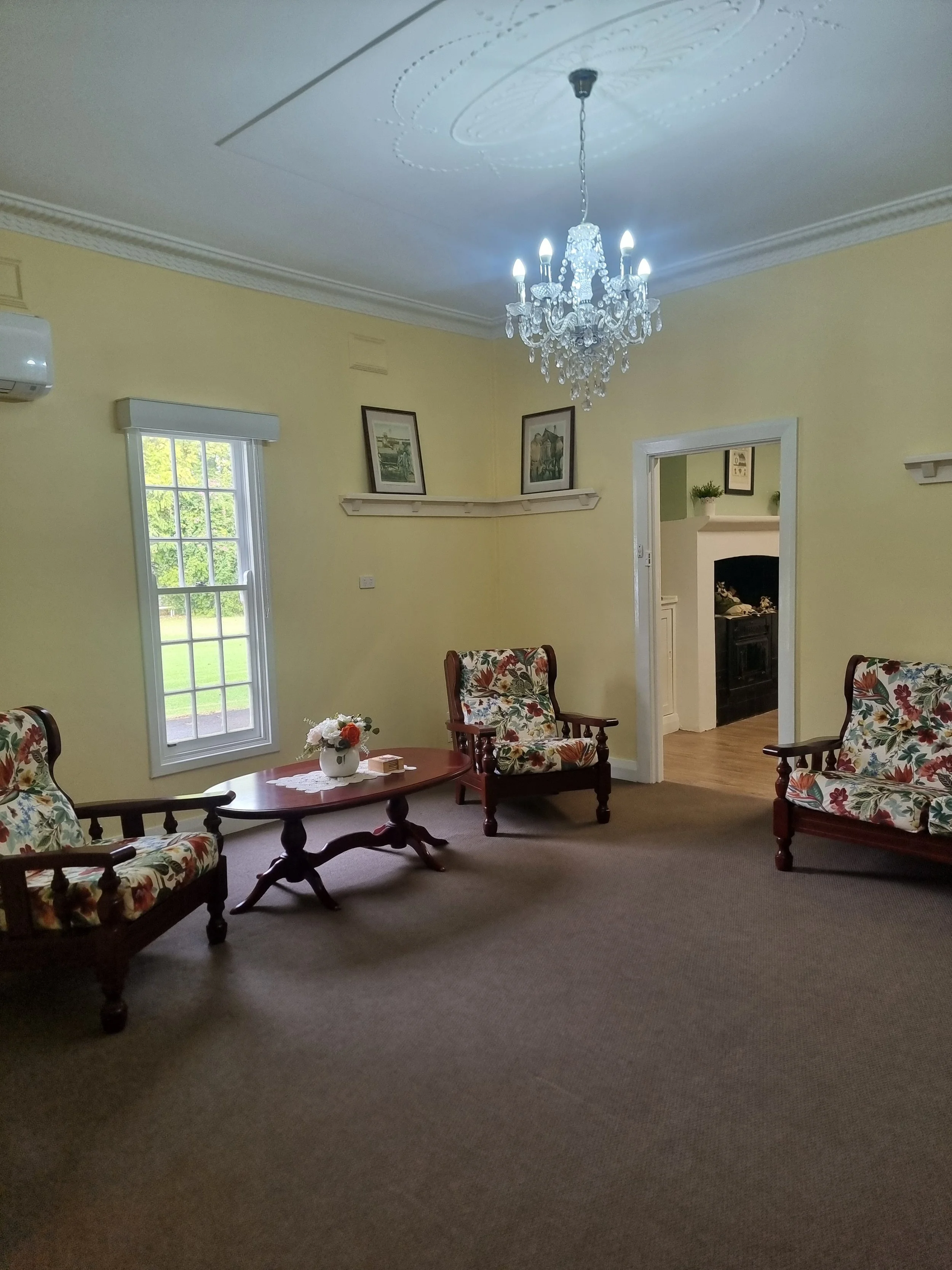 Living room with yellow walls, floral upholstered furniture, a wooden coffee table with a flower arrangement, framed pictures on the wall, and a chandelier hanging from the ceiling.