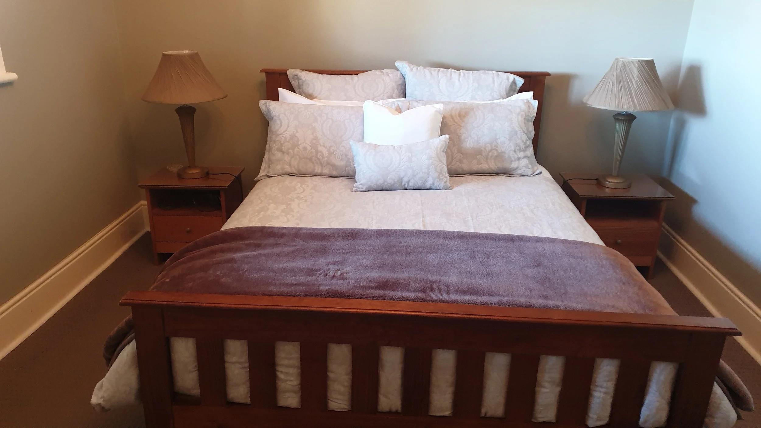 A neatly made bed with multiple white and patterned pillows, a wooden headboard, two bedside tables with lamps, and a brown blanket at the foot of the bed in a cozy bedroom.