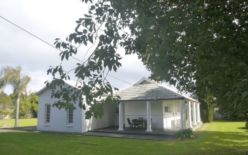 A white house with a porch and outdoor furniture, surrounded by green grass and trees, under a partly cloudy sky.