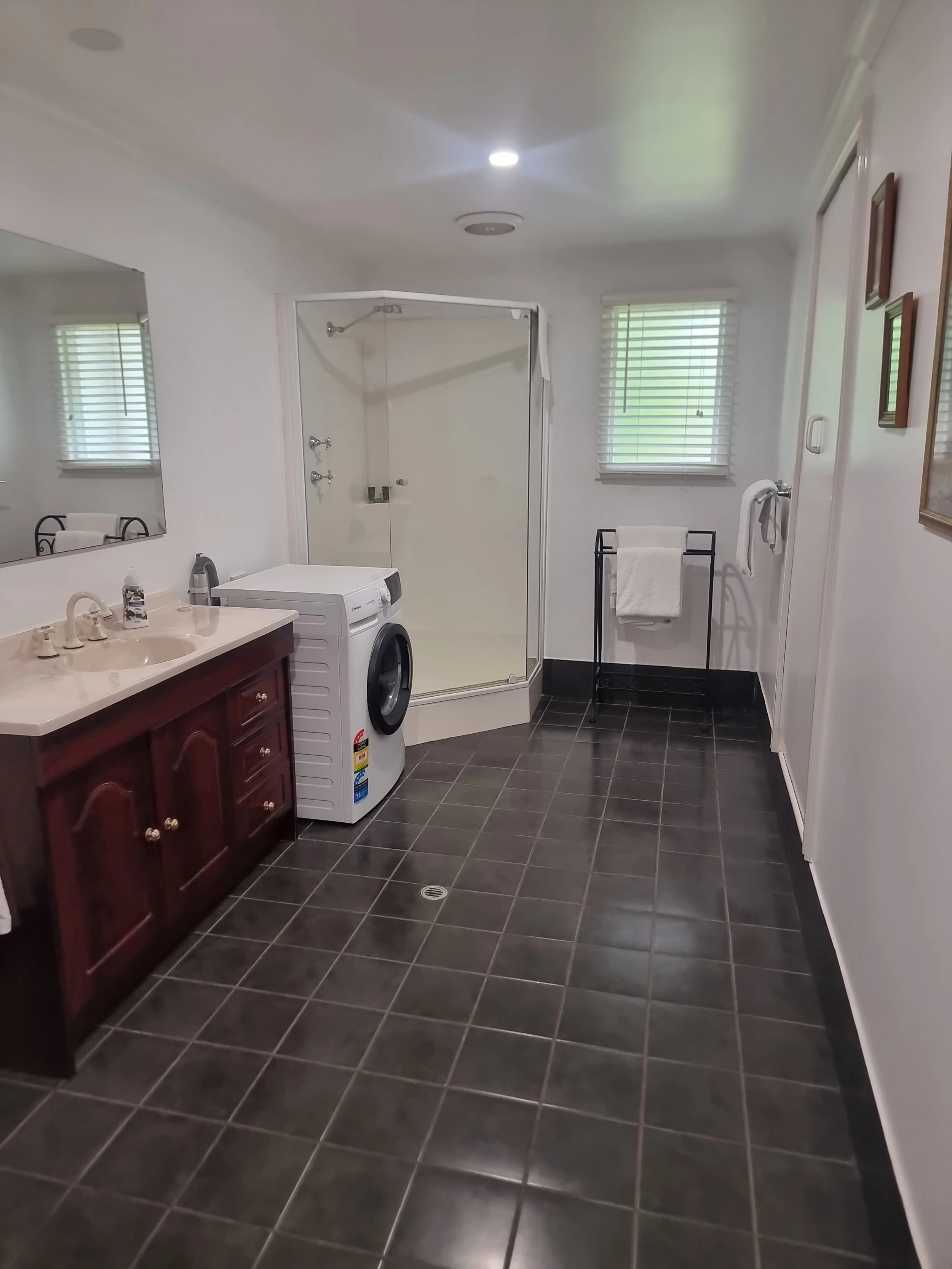 A bathroom with dark tiled floor, white walls, a window with blinds, a mirror above a sink, a washing machine, a shower with glass door, towel rack with towels, and framed pictures on the wall.