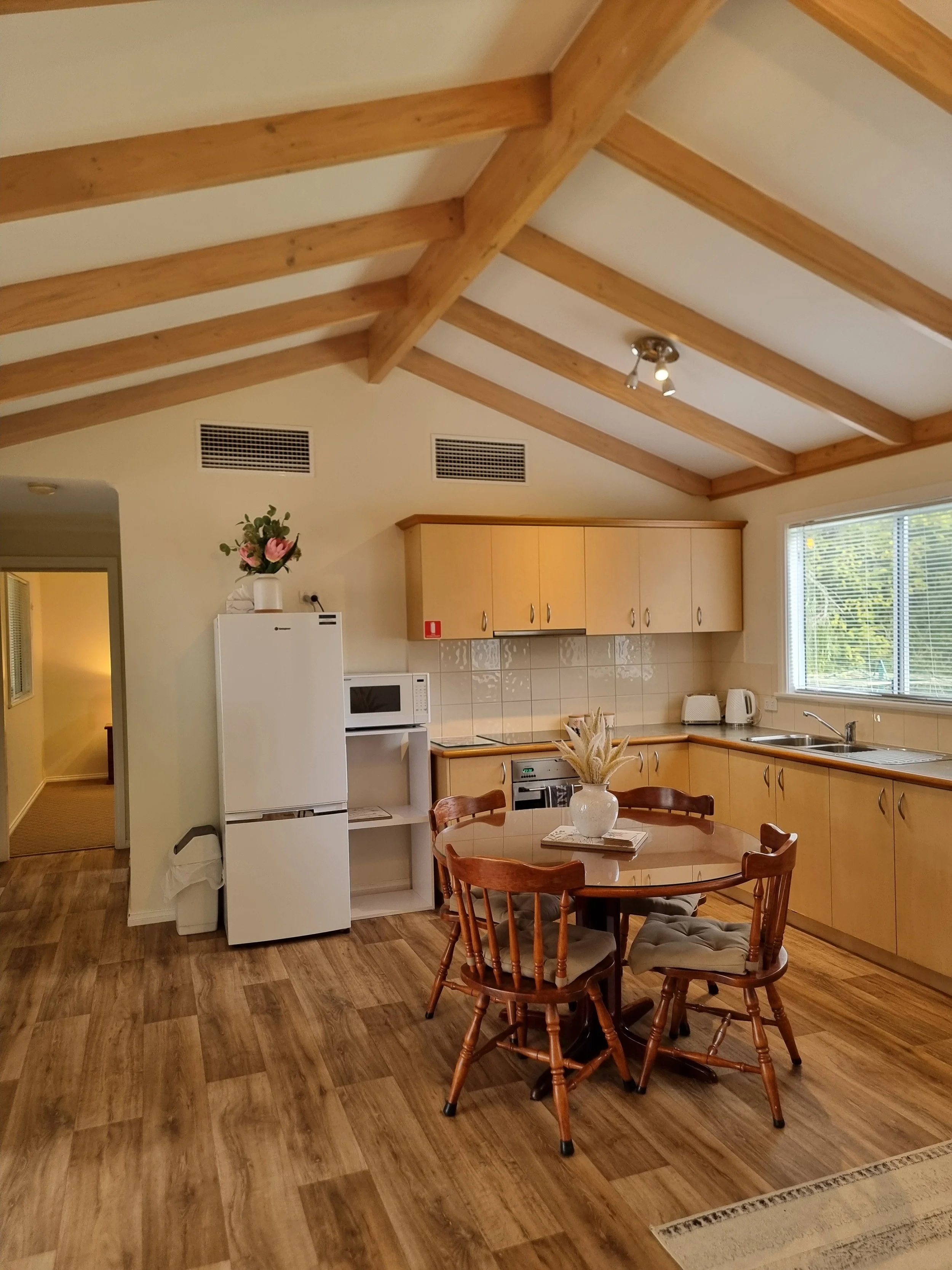 Kitchen with wooden ceiling beams, a white refrigerator, microwave, and countertop appliances, wooden dining table with four chairs, a vase with flowers, and a window with blinds letting in sunlight.