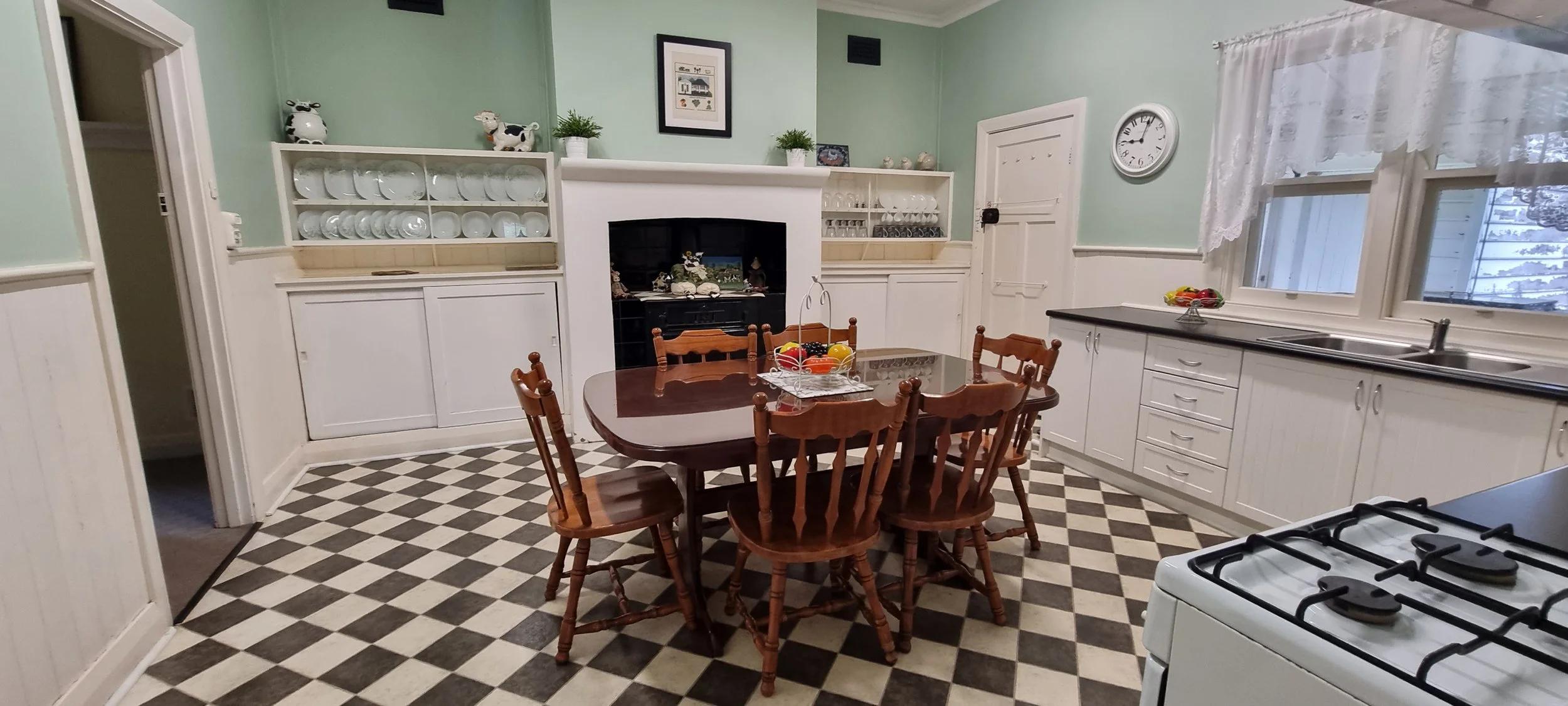 A vintage kitchen with white cabinets, a checkered black and white floor, a wooden dining table with six matching chairs, a fireplace with decorative items, and a window with lace curtains overlooking the outside.