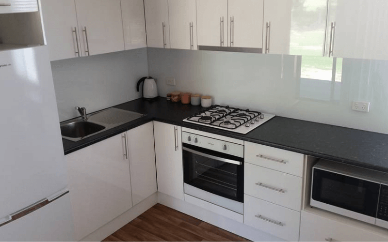 Modern kitchen with white cabinets, black countertops, a stove, microwave, refrigerator, small sink, kettle, and jars, with a window letting in natural light.