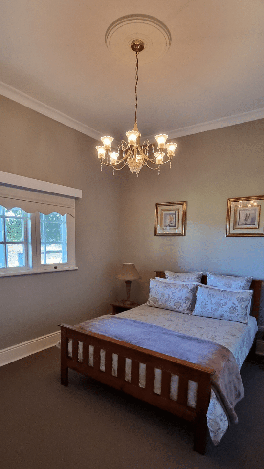 Bedroom with a wooden bed, patterned bedding, a nightstand with a lamp, framed art on the wall, a window with a white valance, and a chandelier hanging from the ceiling.