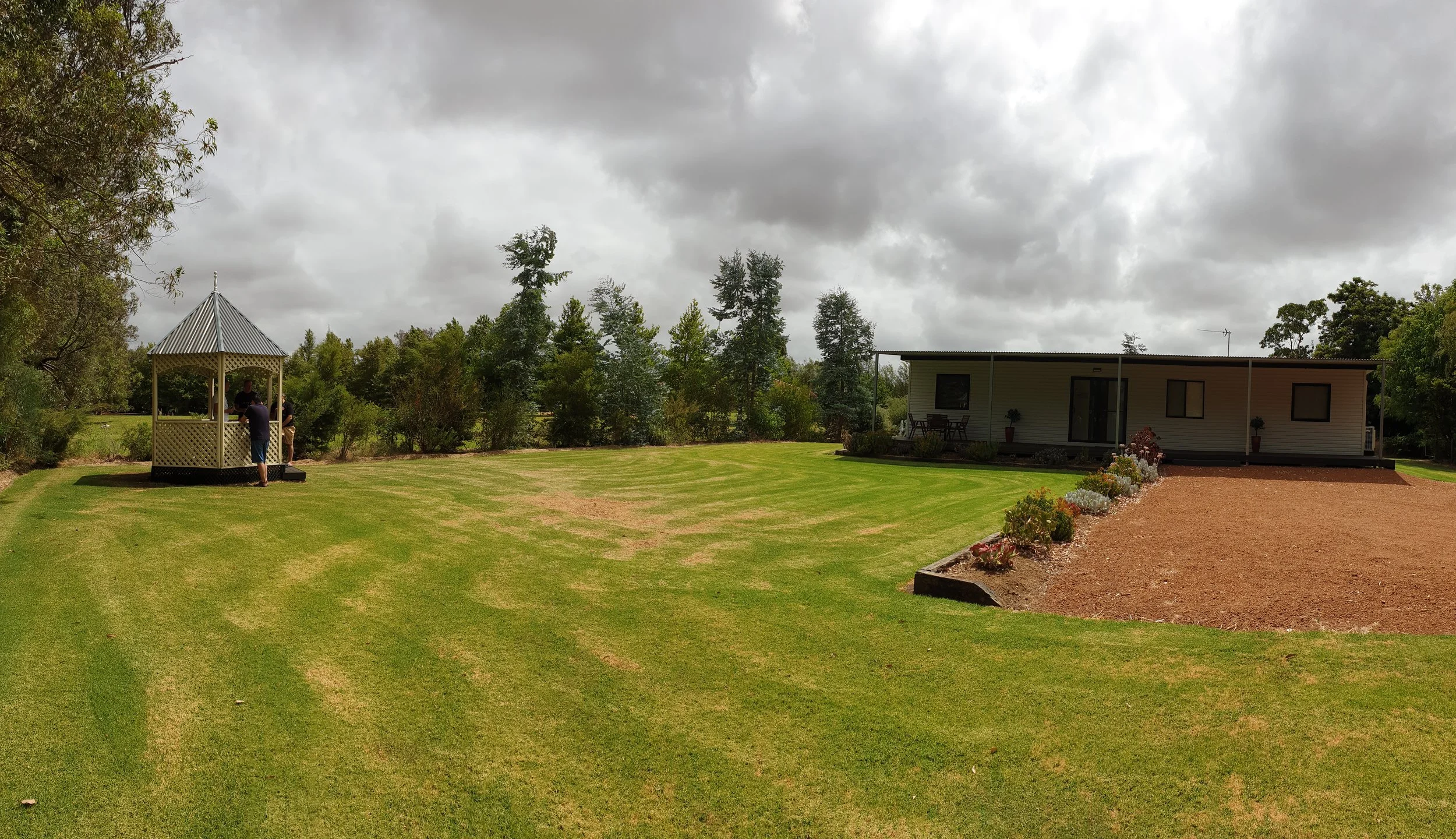 A backyard with a green lawn, small white gazebo with a gray roof where a person is standing, a modern single-story house with a deck, potted plants, and garden beds, surrounded by trees under a cloudy sky.
