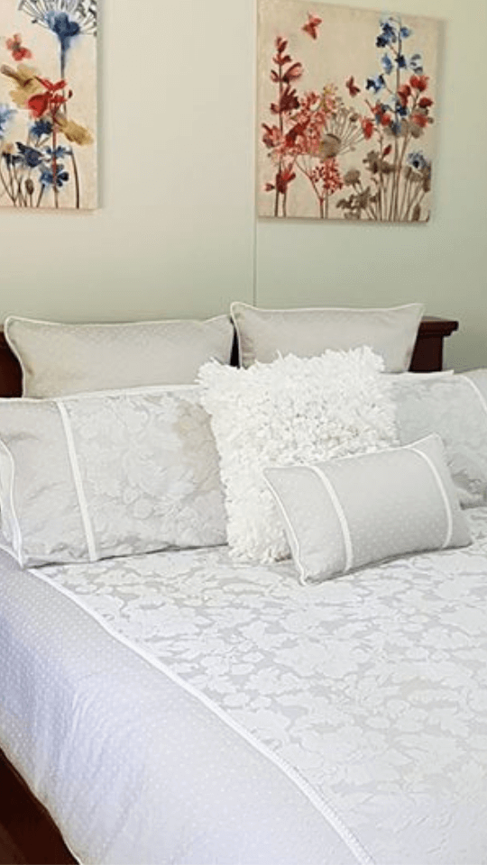 A neatly made bed with white floral-patterned bedding, adorned with multiple white pillows and a fluffy white decorative pillow, with floral artwork hanging on the light-colored wall above.