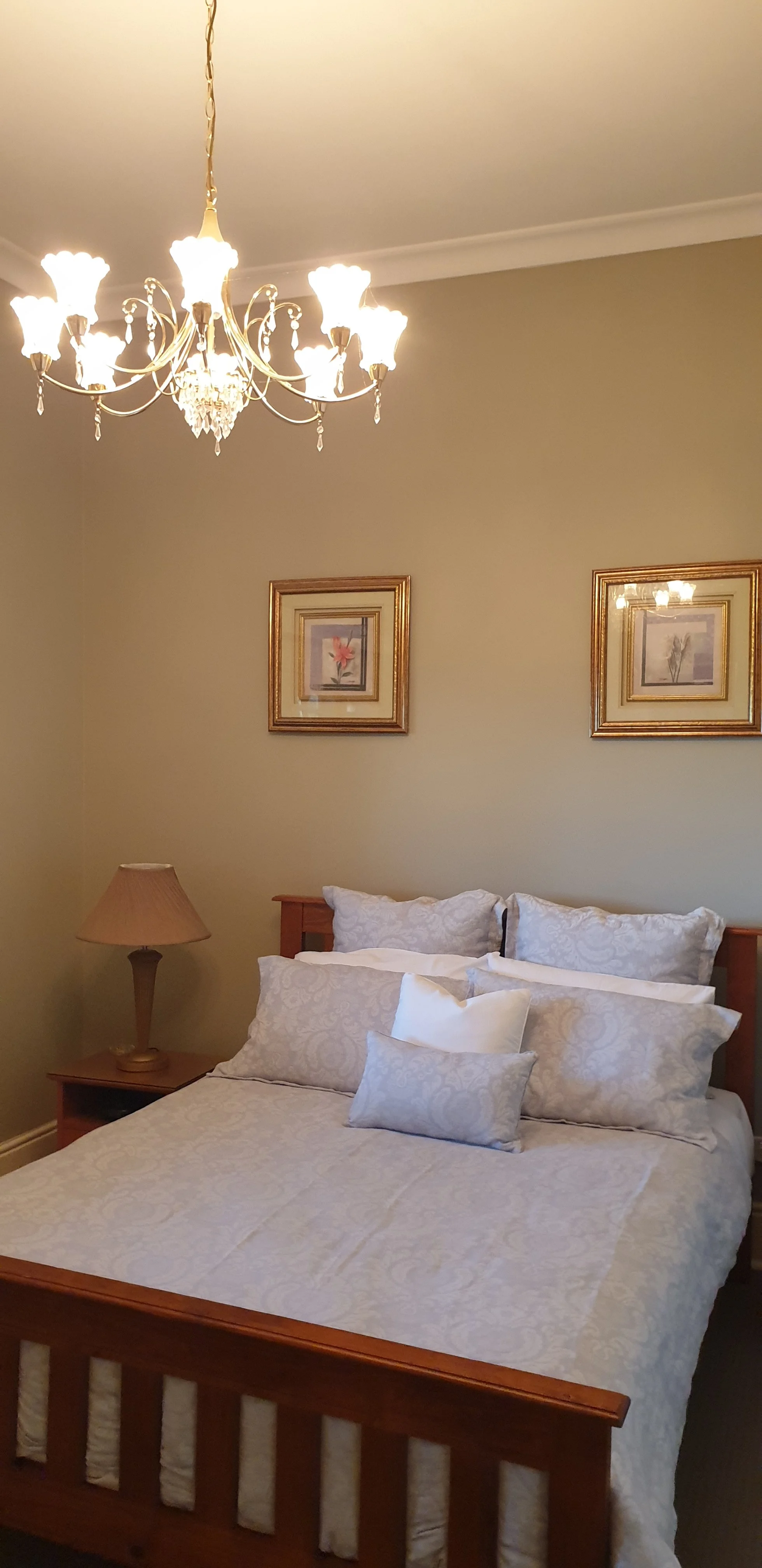 A bedroom with a wooden bed frame, a white quilt, several pillows, a wooden nightstand with a beige lamp, two framed pictures of flowers on the wall, and a chandelier hanging from the ceiling.