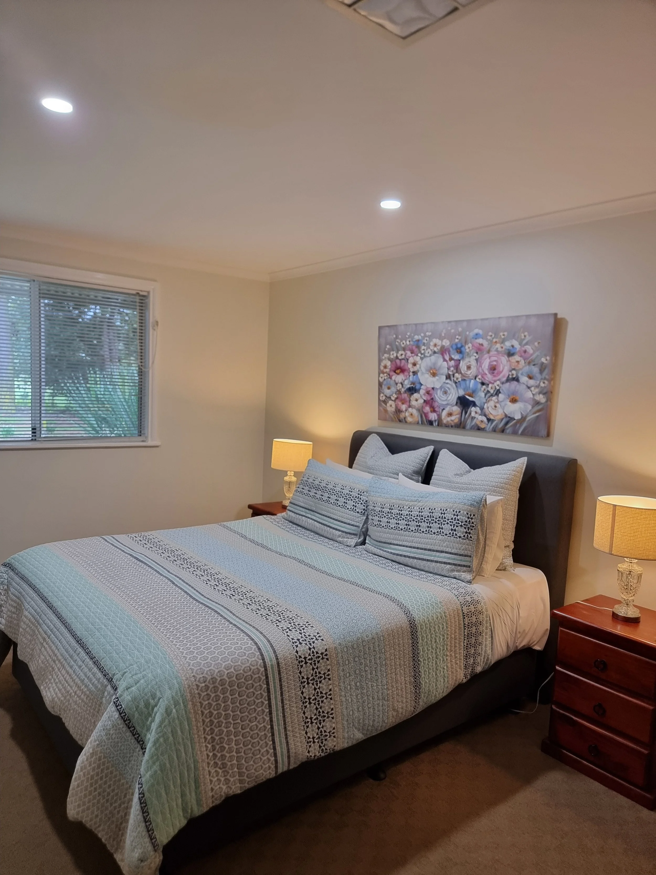 A bedroom with a bed covered in a white and blue patterned quilt, two pillows, and a headboard. Two wooden nightstands with lamps are on either side of the bed. A large painting of flowers hangs above the headboard. A window with blinds is on the left wall, allowing natural light into the room.