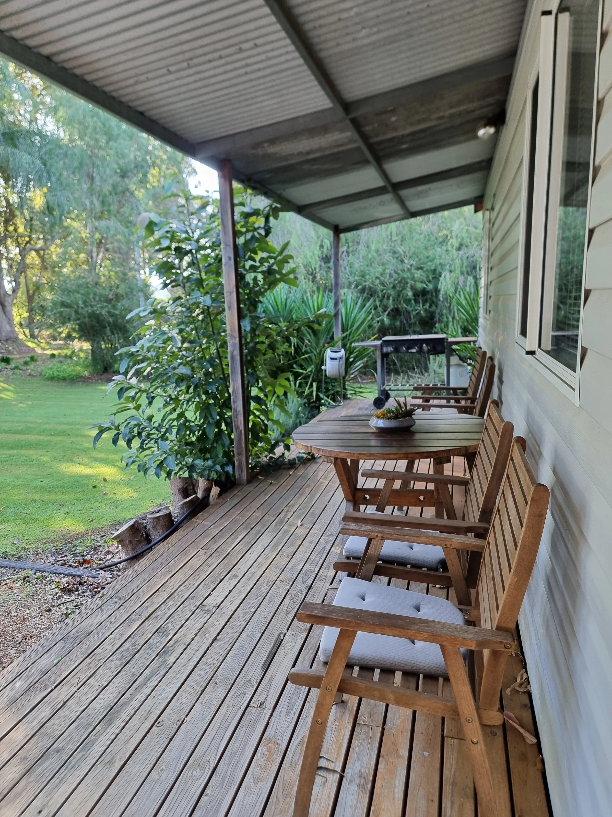A wooden porch with outdoor chairs and a table, surrounded by lush green trees and plants, with a metal roof overhead.
